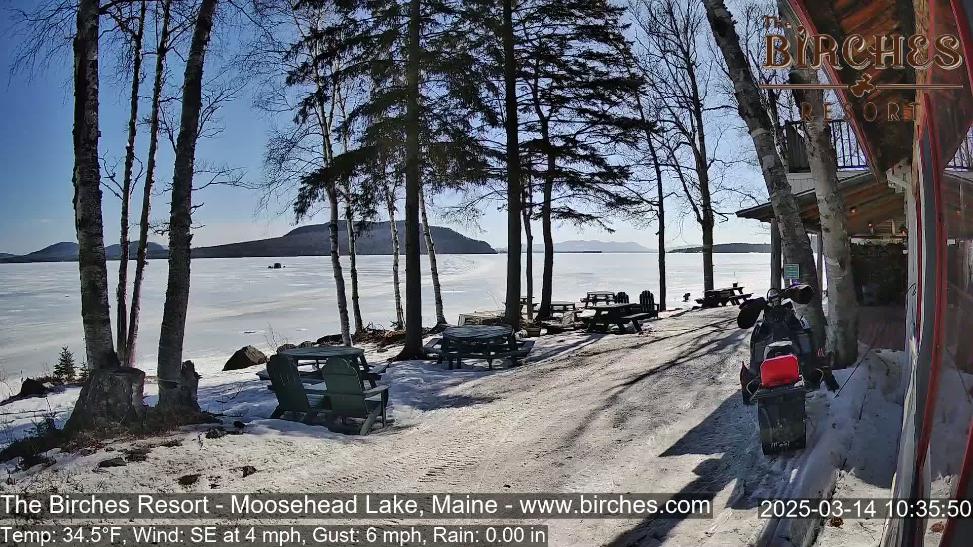 A snow-covered lakeside area with picnic tables, a snowmobile, and trees, under a sunny sky with a partly frozen lake in the background.