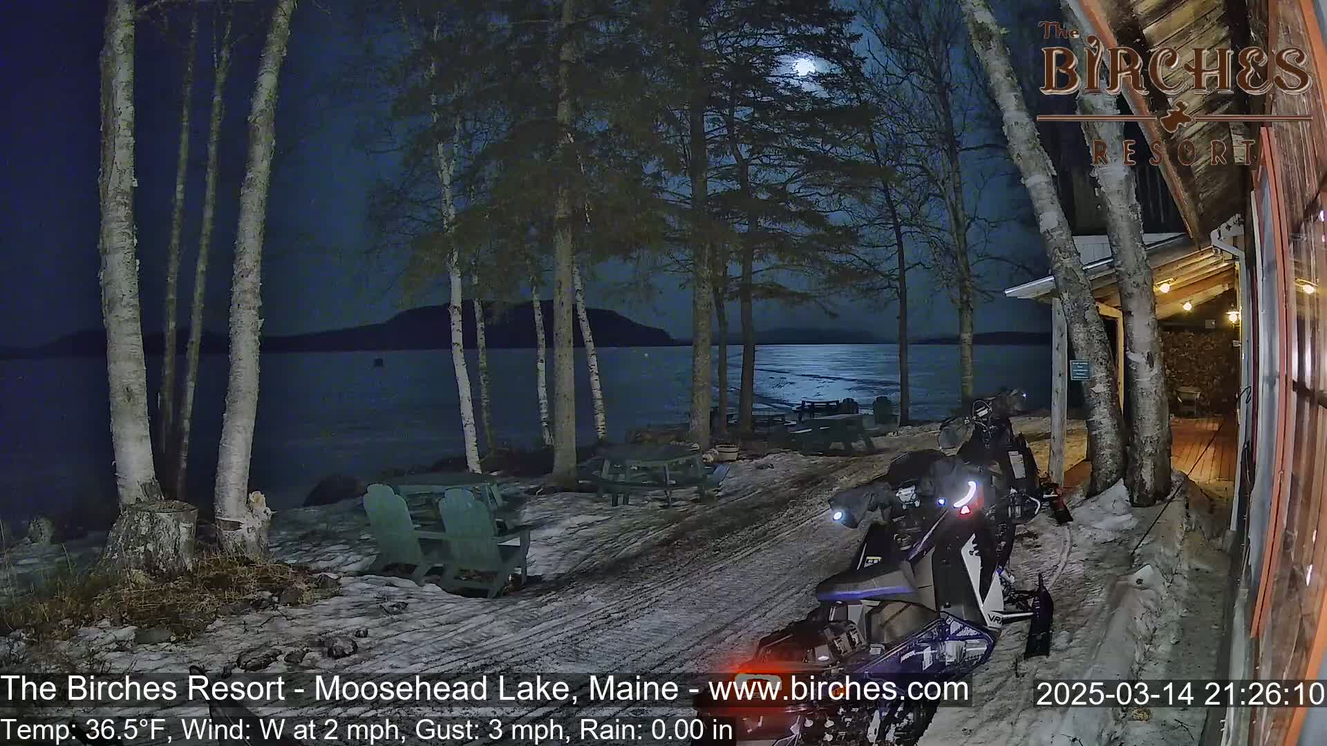 A snow-covered lakeside area at night under a full moon, featuring a snowmobile parked near a building, with calm water visible beyond some birch trees.