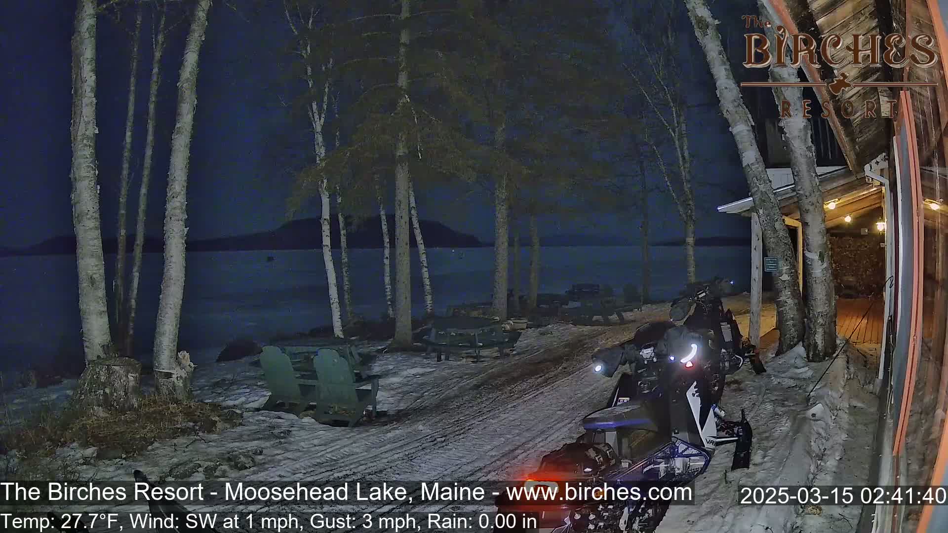 A snowmobile is parked near a lakeside resort at night under a dark sky, with a light dusting of snow on the ground.