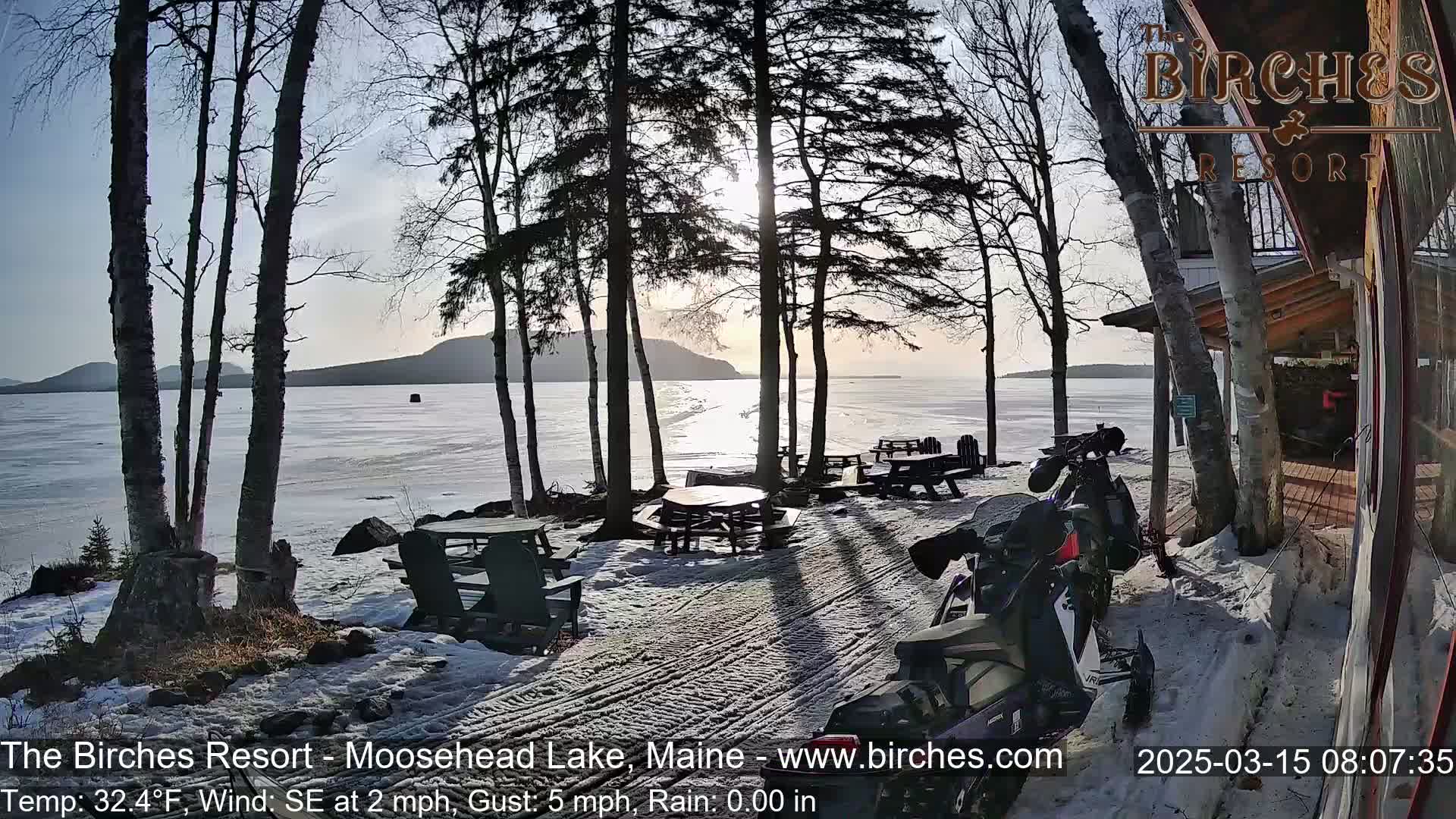 A snow-covered lakeside area with picnic tables, Adirondack chairs, and a snowmobile parked near a building, under a partly sunny sky.