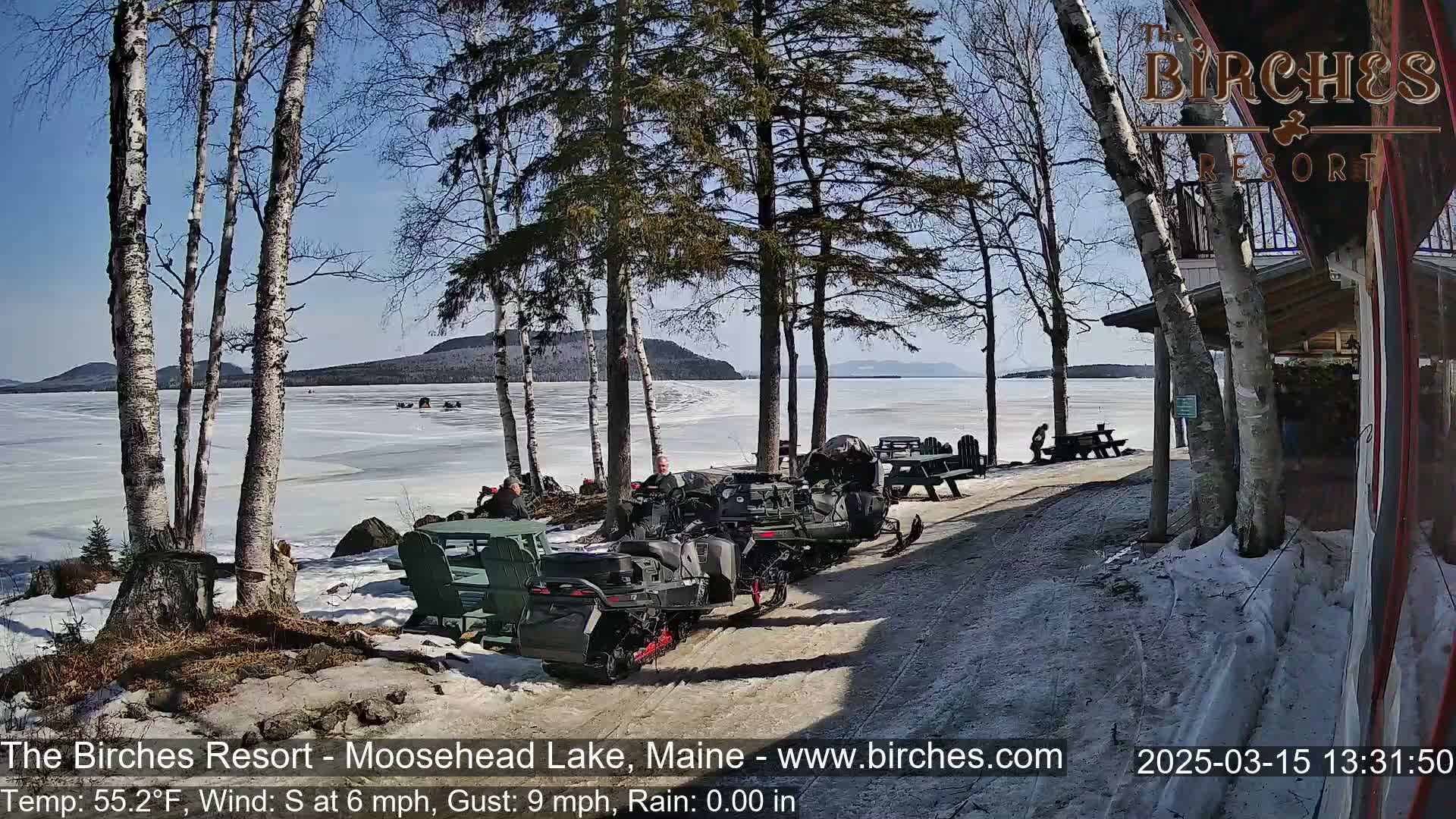 Several snowmobiles are parked near a lakeside resort on a sunny day with snow on the ground and a partially frozen lake in the background.