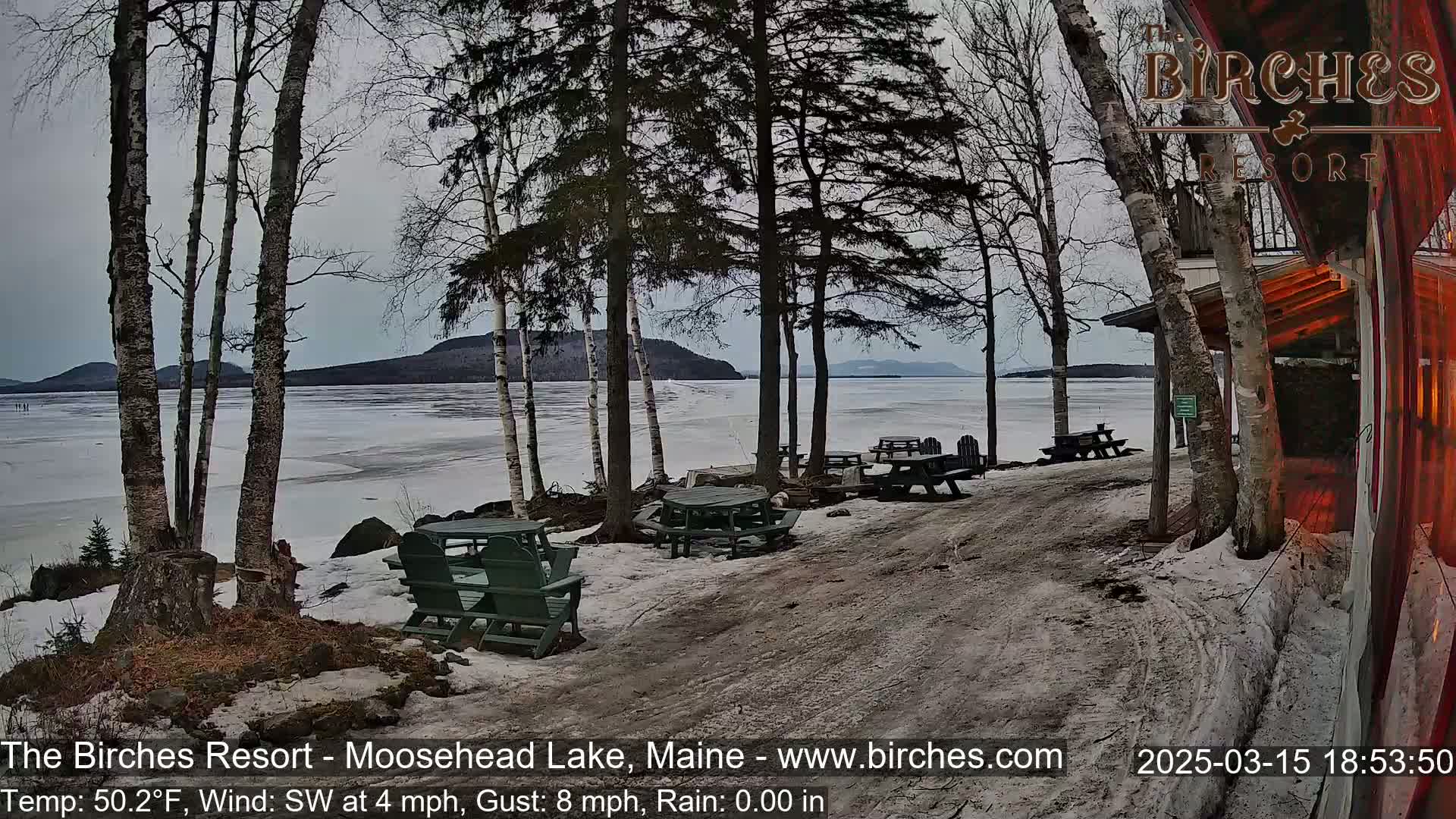 A partially frozen lake is viewed from a lakeside resort area with several empty picnic tables and chairs on a partly snowy, overcast day.