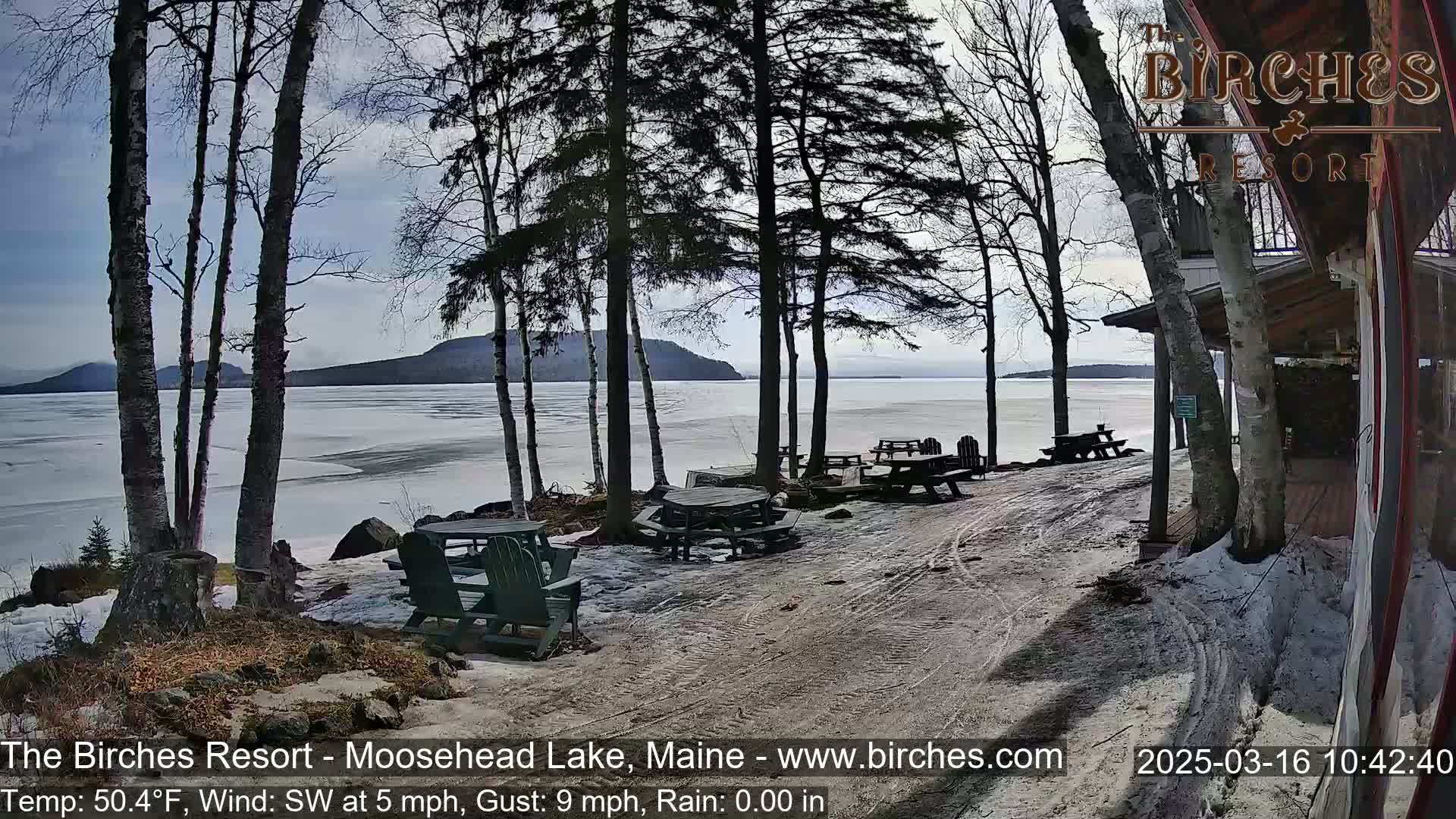A partly sunny day with a light southwest wind reveals a partially frozen lake with picnic tables and chairs near a lakeside building, surrounded by bare trees.