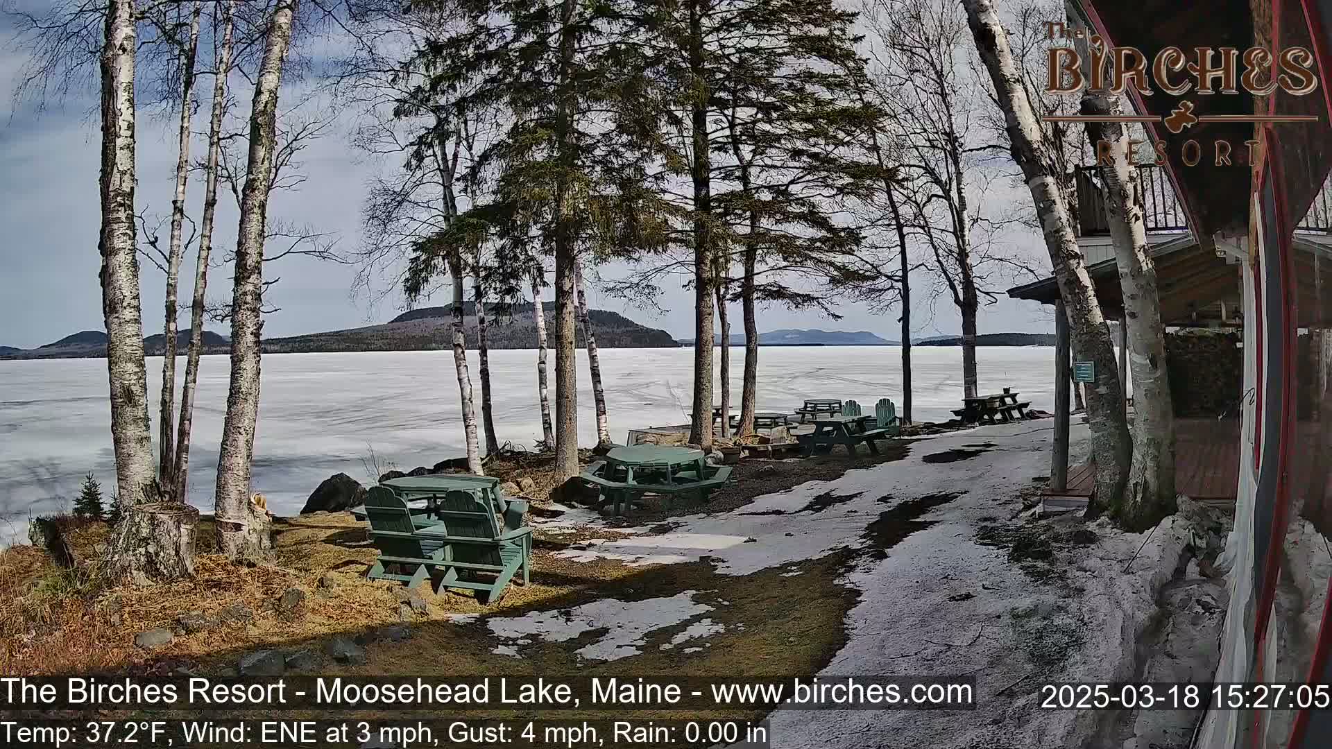 A partially frozen lake is visible beyond a lakeside patio with picnic tables and chairs, under a partly sunny sky with patchy snow on the ground.