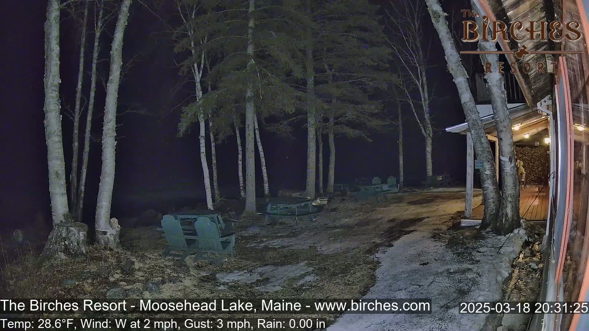 A nighttime outdoor scene shows several empty Adirondack chairs near a building under a dark sky with a slight west wind.