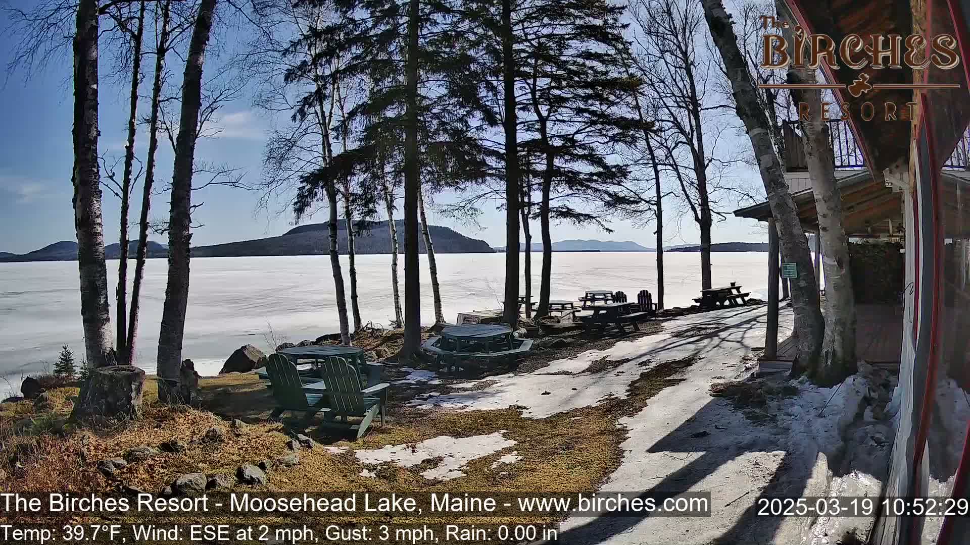 A partially frozen lake is visible beyond a lakeside patio area with picnic tables and chairs, under a mostly sunny sky.