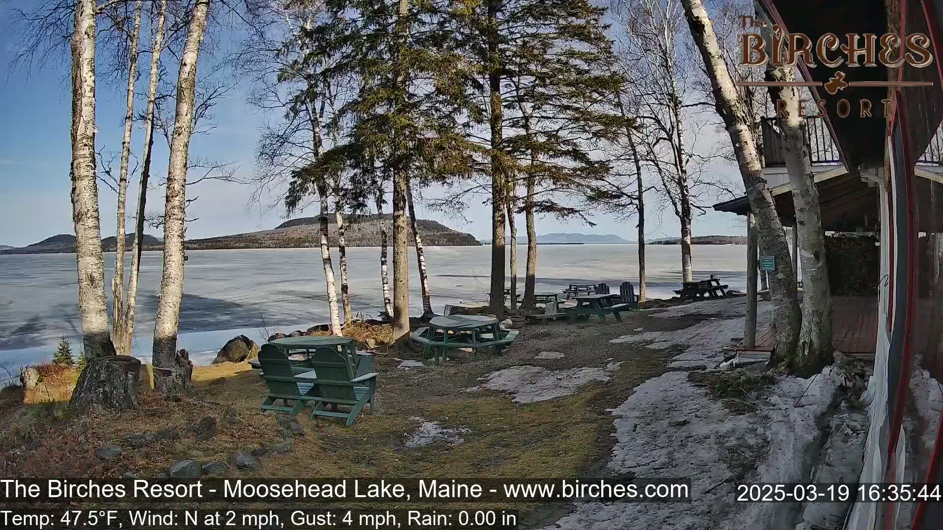 A partly sunny view of a partially frozen lake with picnic tables and chairs near a lakeside building, surrounded by birch and evergreen trees.