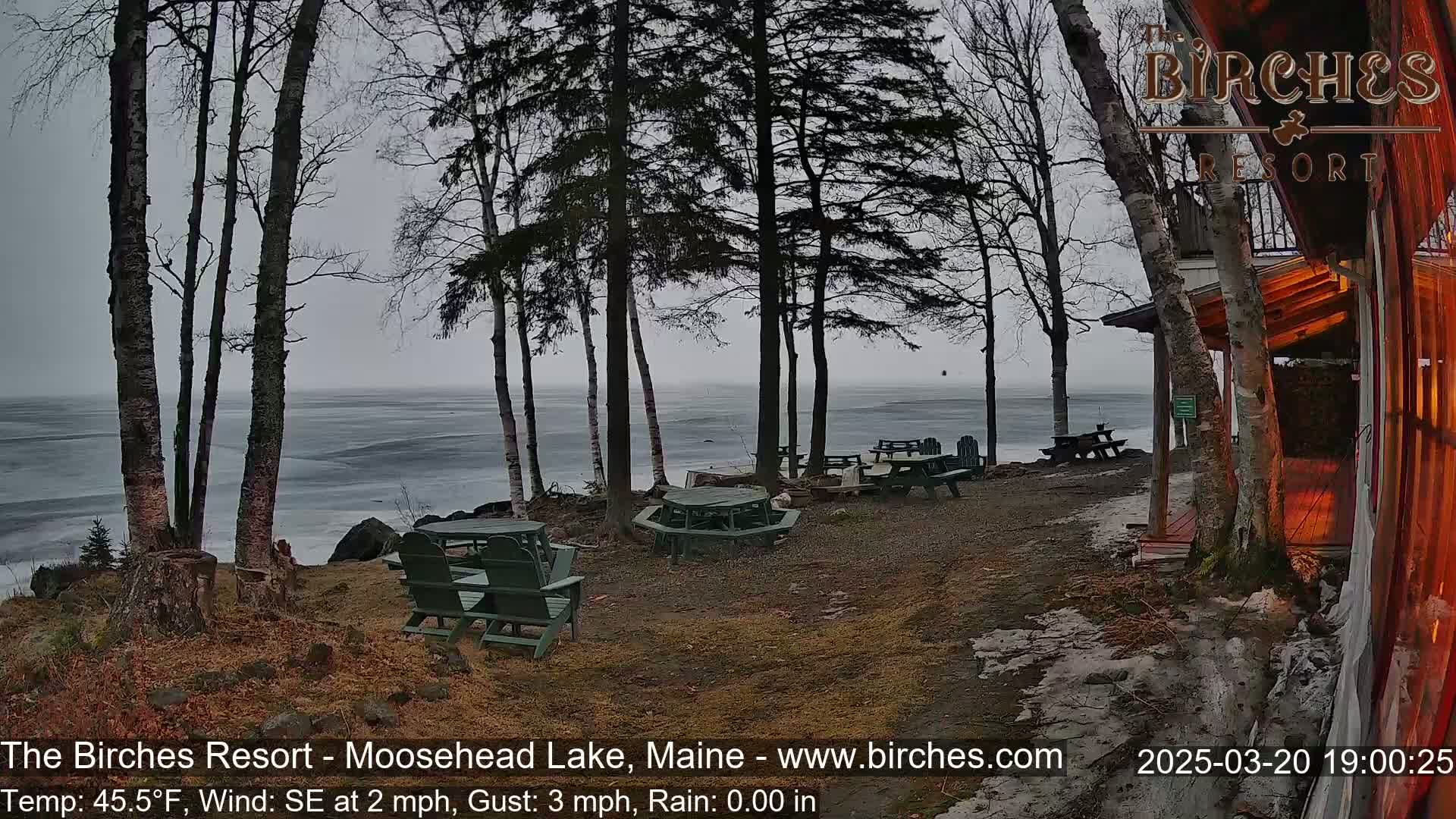 A partially frozen lake is visible from a lakeside patio area with several empty picnic tables and chairs, under an overcast sky with a light southeast wind.