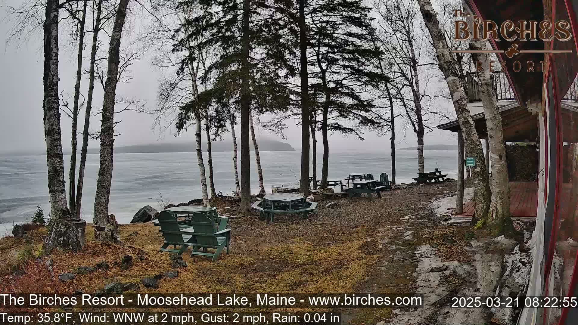 A lakeside patio with picnic tables and chairs is partially covered in snow under a rainy, overcast sky.