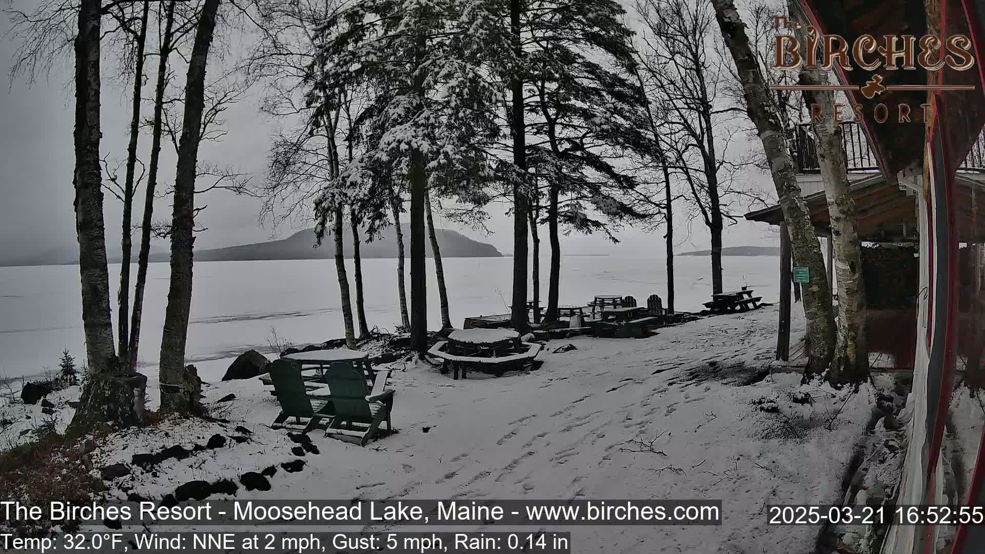 A snow-covered lakeside patio with picnic tables and chairs overlooks a partially frozen lake under a gray sky.