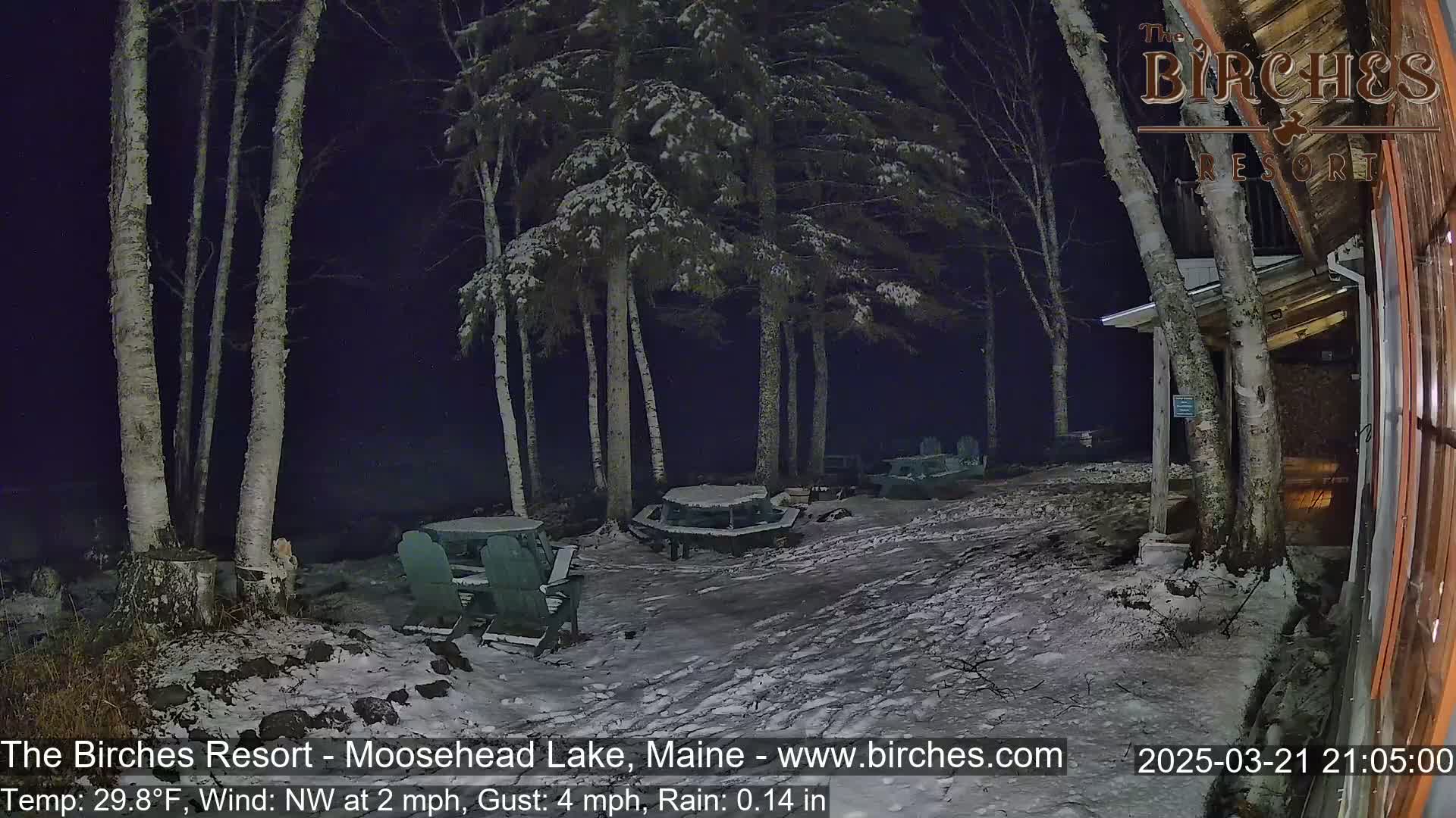 A snow-covered outdoor area at night, illuminated by faint light, shows several picnic tables and chairs near a building under a dark sky with a light snowfall.