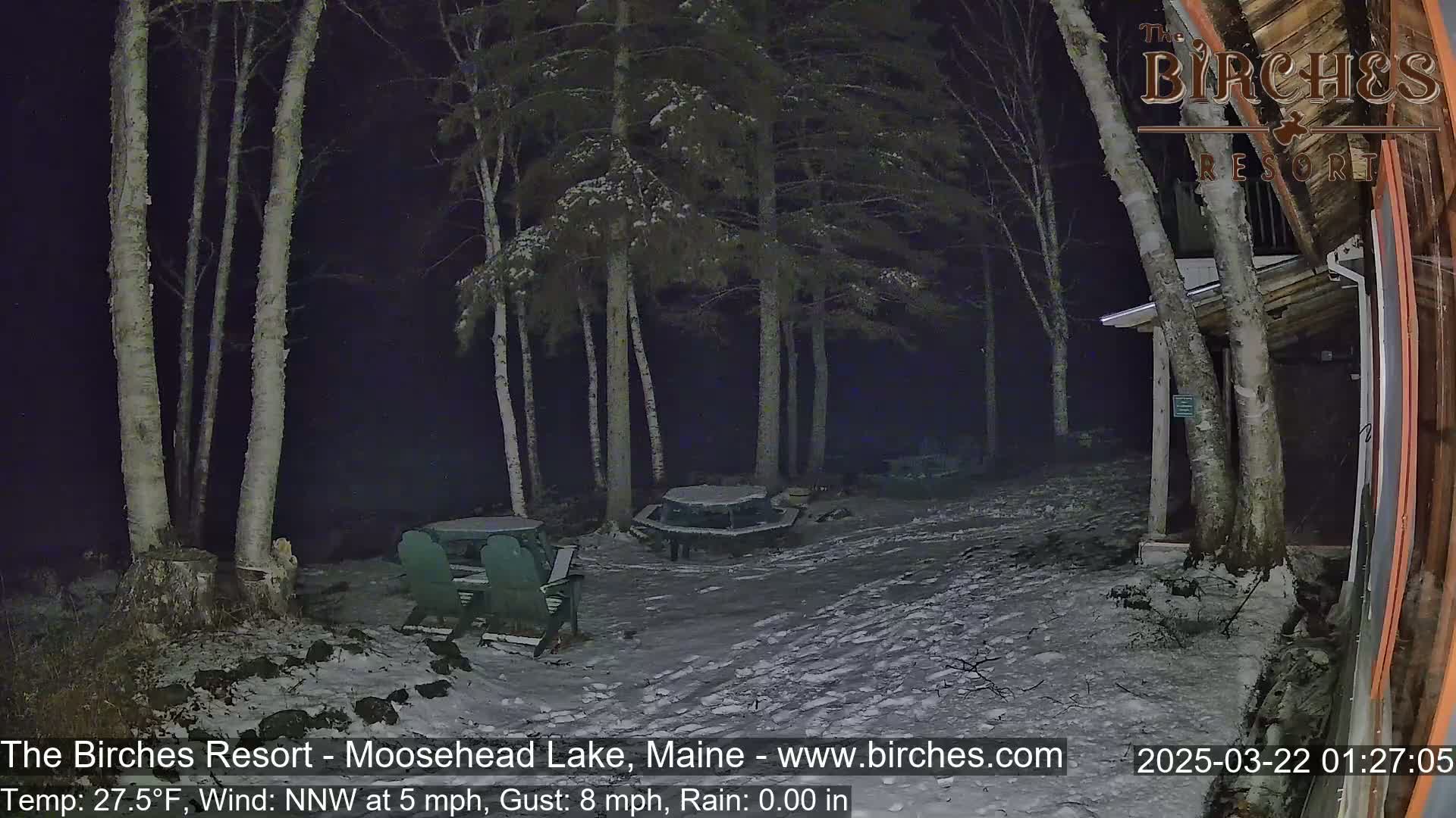 A snow-covered outdoor area at night, partially illuminated, shows a picnic table and chairs near several birch trees and a building under a dark, cloudy sky.