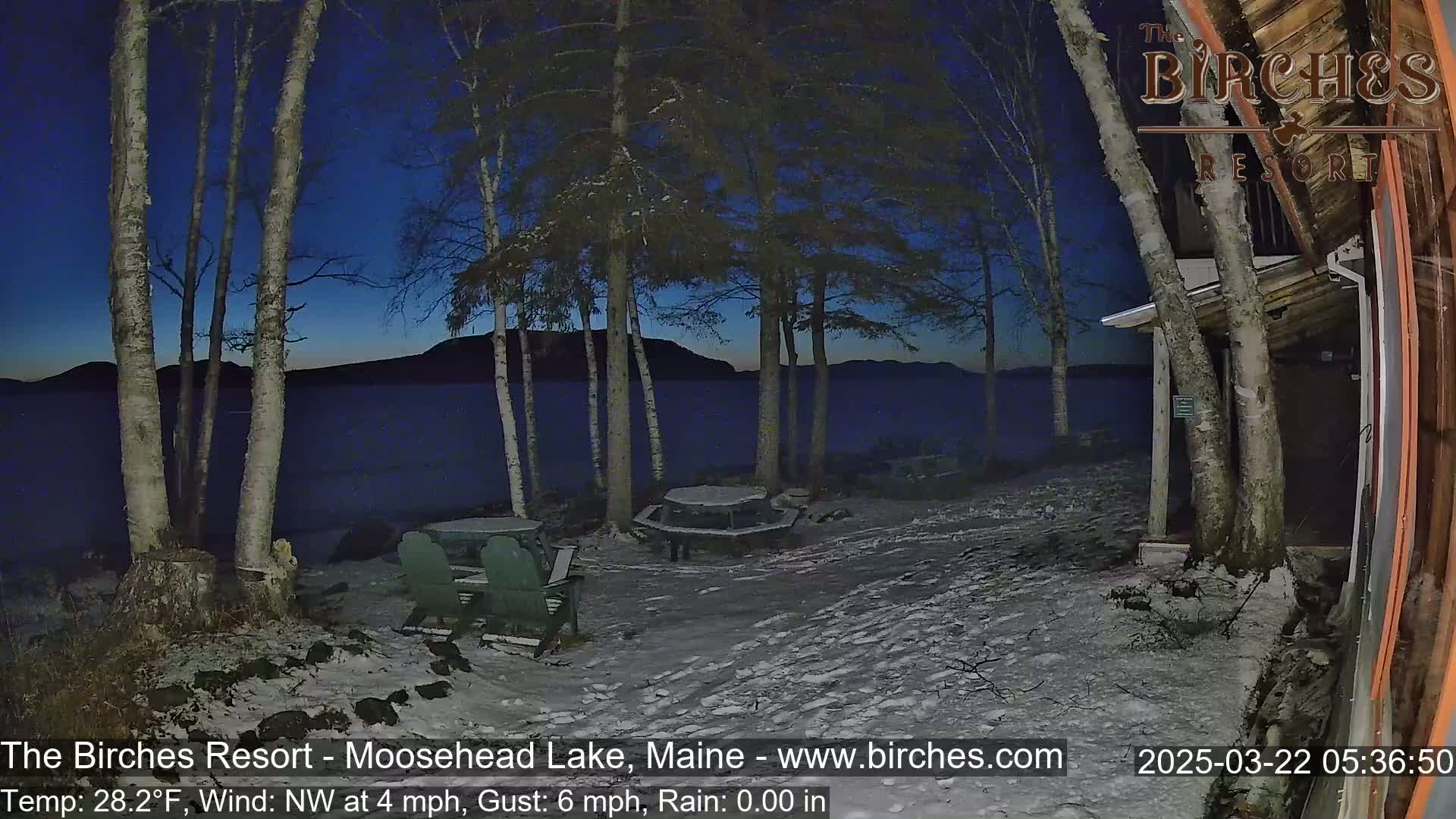 A snow-covered lakeside area at night, with several birch trees, picnic tables, and Adirondack chairs, under a dark blue sky showing a hint of pre-dawn light.