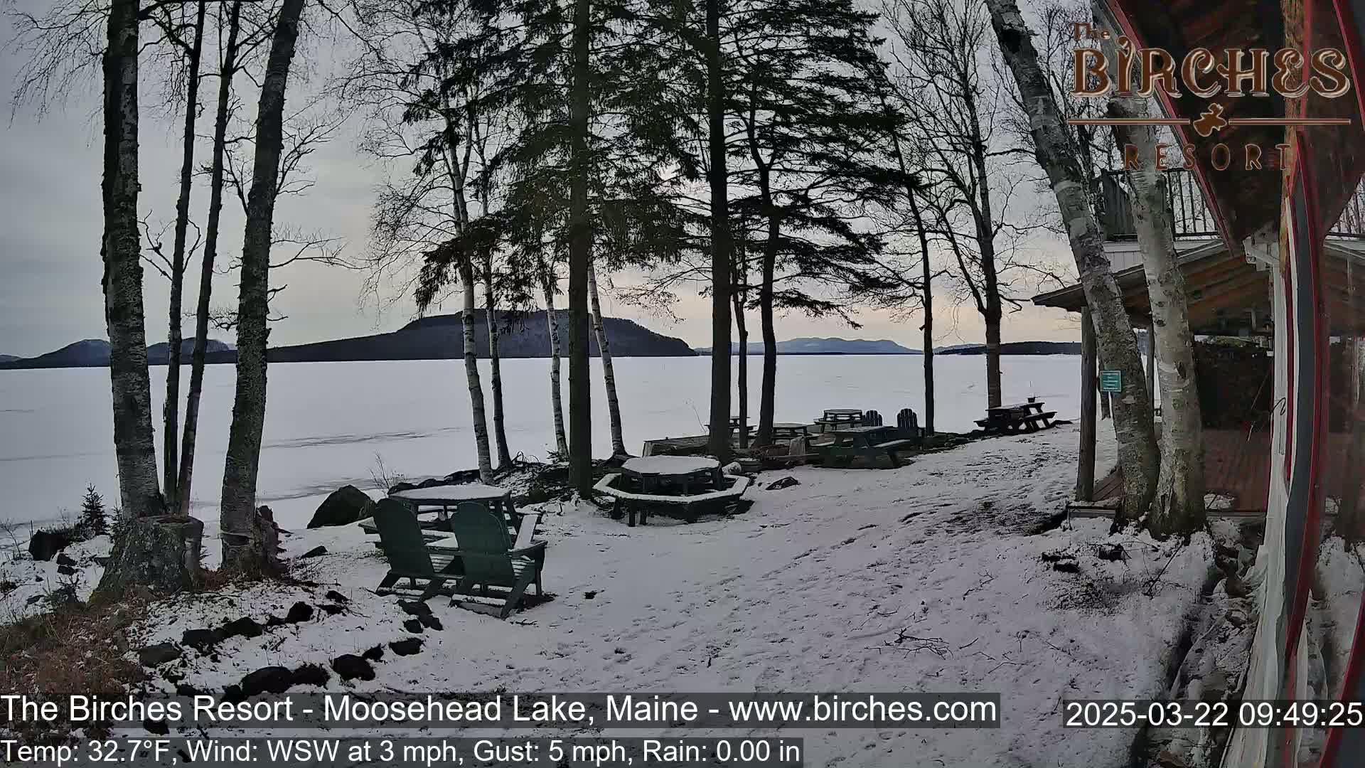 A snow-covered lakeside patio with picnic tables and chairs overlooks a frozen lake and distant hills under a cloudy sky.
