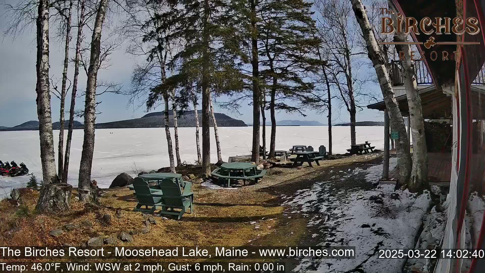 A partly sunny view of a frozen lake with snow-covered ground and picnic tables near a lakeside building, with snowmobiles visible in the distance.