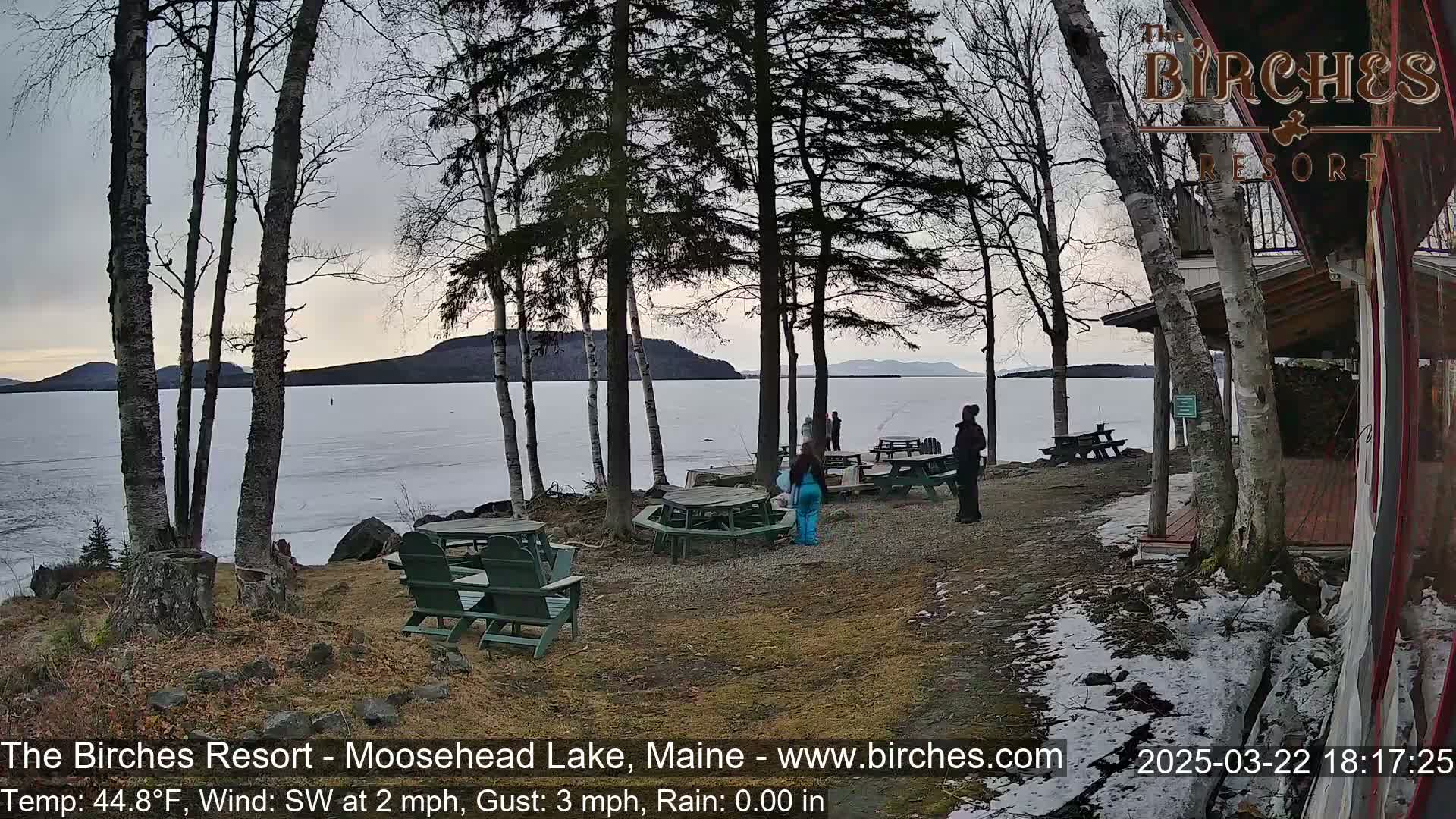 A partially frozen lake is seen from a lakeside resort with several people gathered near picnic tables under a cloudy sky.