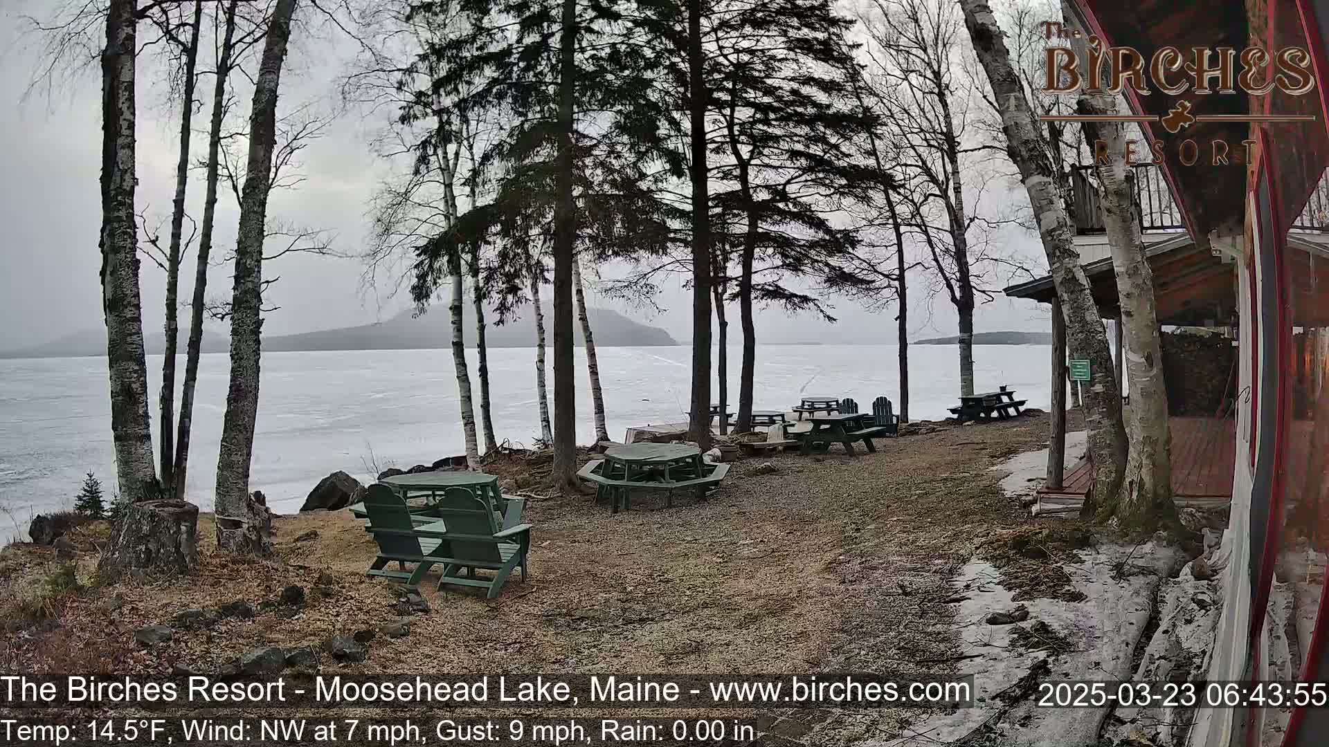 A partly frozen lake is visible from a lakeside patio area with several picnic tables and chairs under a gray, overcast sky.