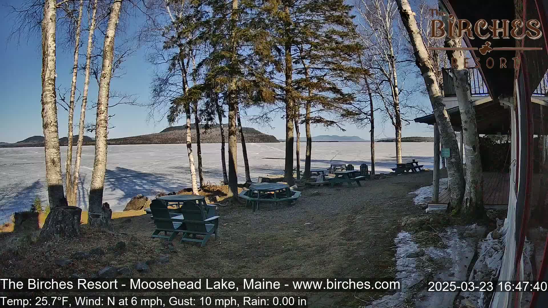 A sunny, partially frozen lake is seen from a lakeside patio with picnic tables and chairs under bare trees on a partly sunny day with a light north wind.