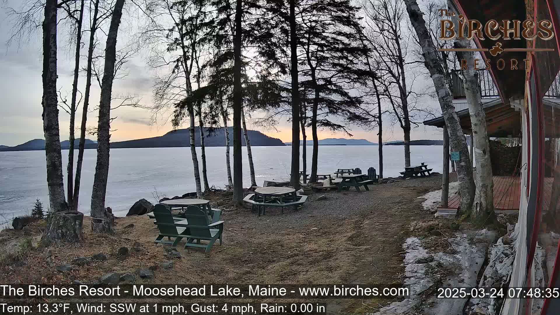Several picnic tables and chairs sit on a lakeside patio near bare trees at sunset, with a partly frozen lake and distant hills visible in the background, under a mostly clear sky.