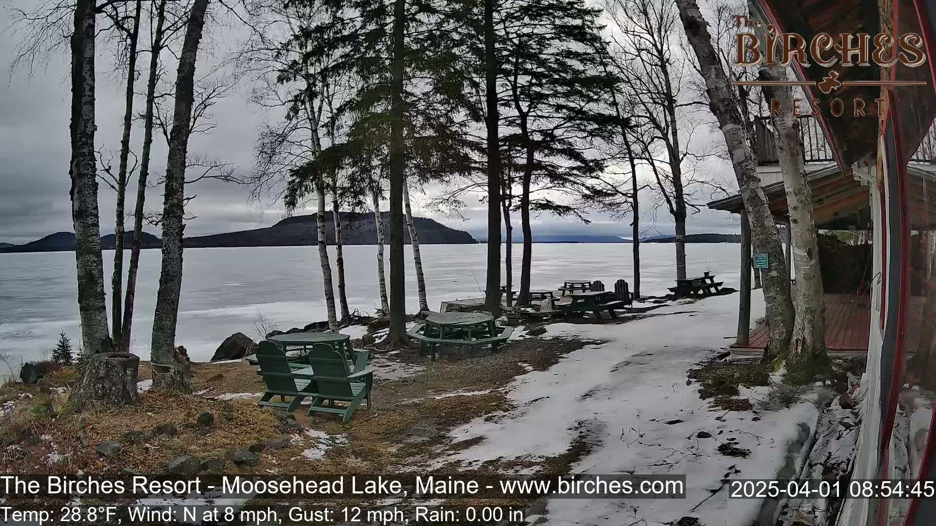 Partially frozen lake with snow-covered ground and picnic tables near a lakeside building under an overcast sky.