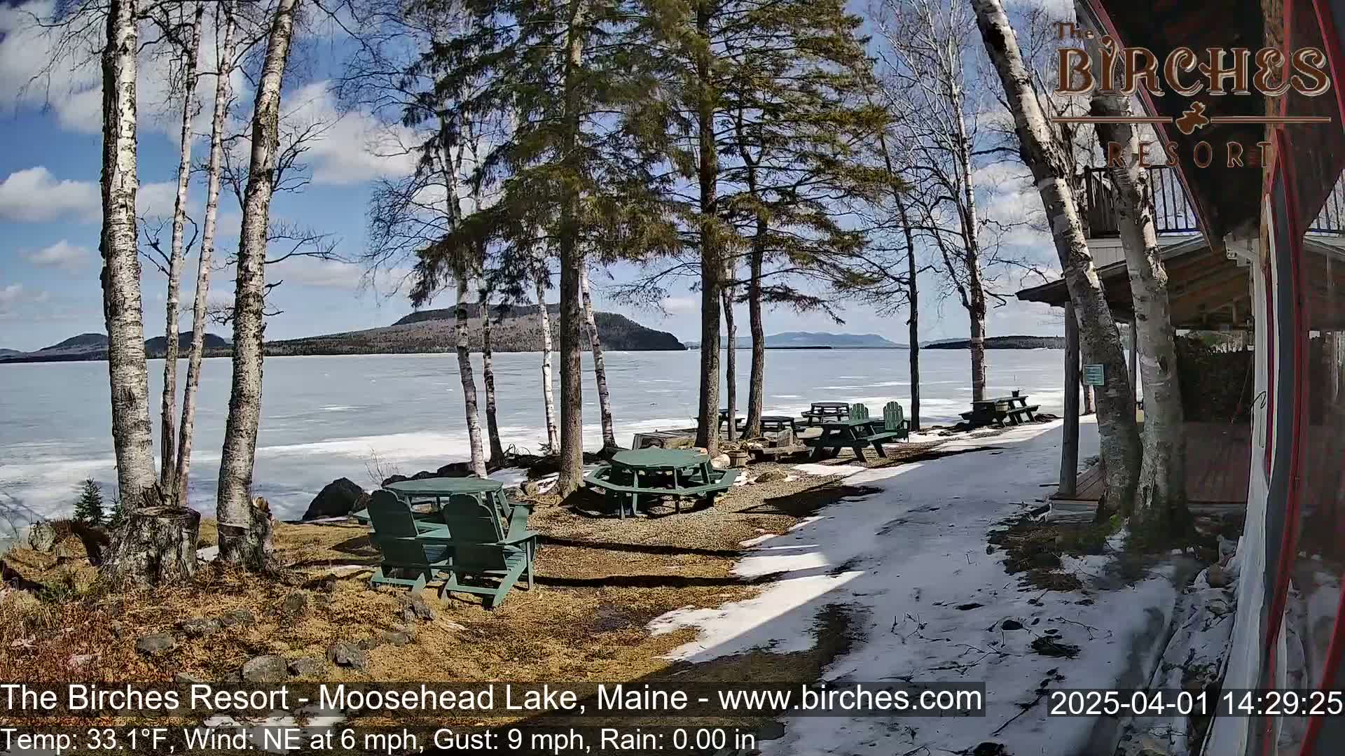 A partly sunny lakeside scene with partially frozen water, picnic tables, and chairs near a building, under a partly cloudy sky.