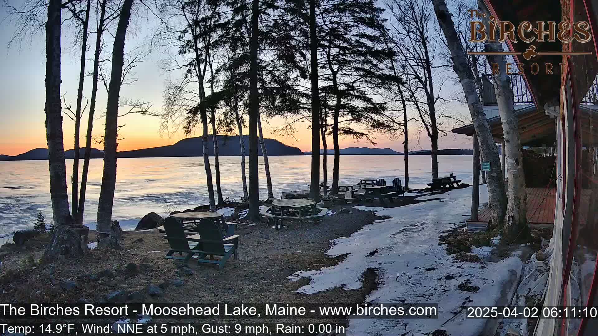 A lakeside patio with picnic tables and chairs partially covered in snow at sunset, overlooking a partially frozen lake under a partly cloudy sky.