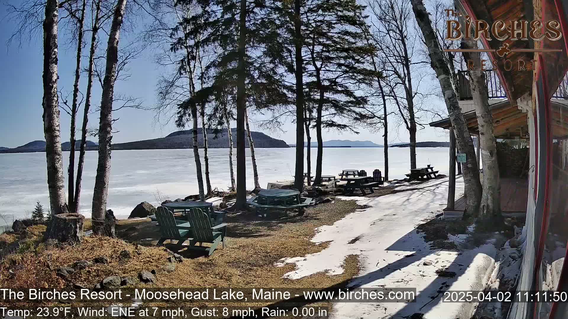 A partly sunny day with a light wind shows a partially frozen lake viewed from a lakeside patio with several picnic tables and chairs, surrounded by birch trees and a building.