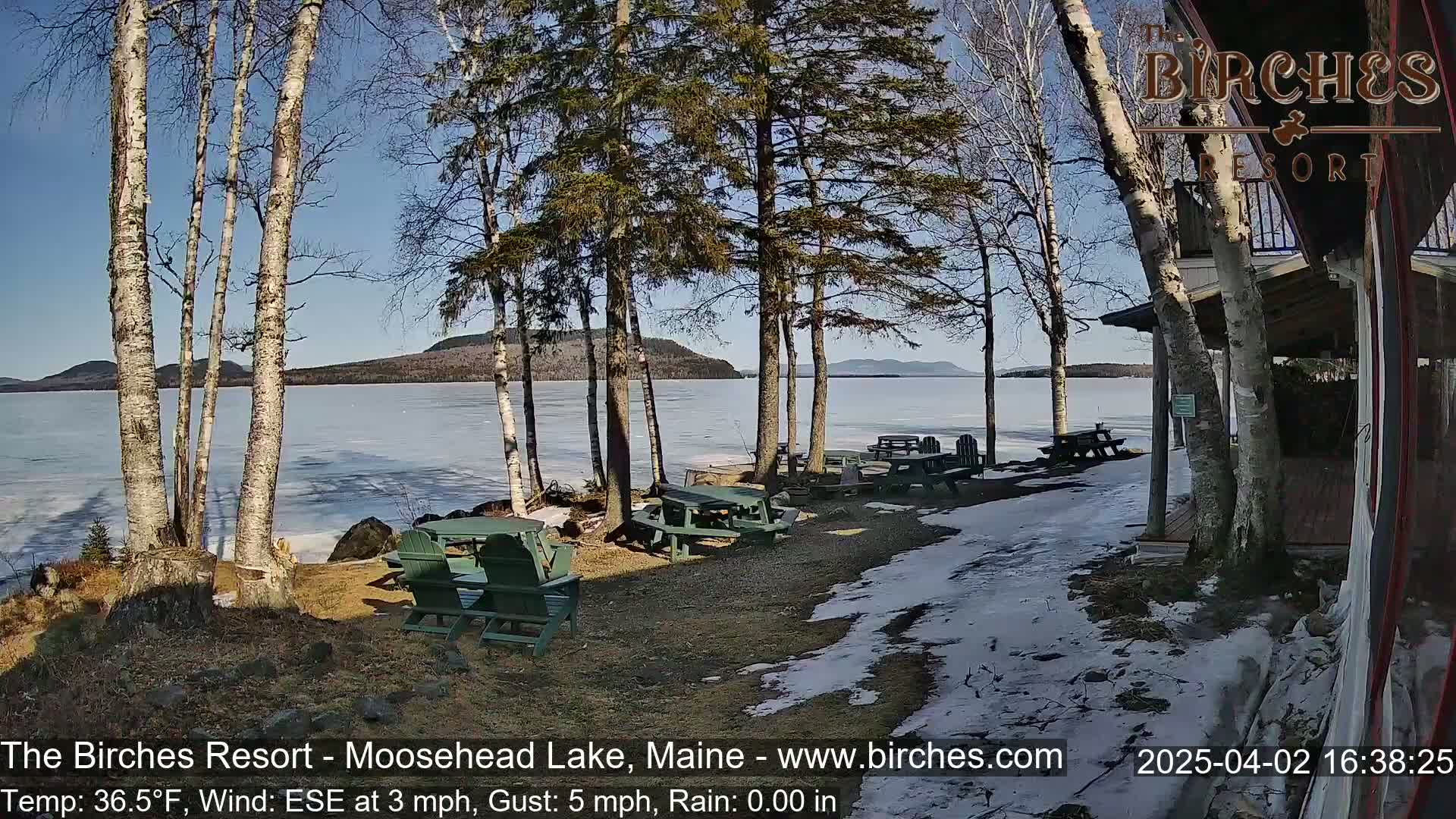 A partly sunny lakeside scene with partially frozen water, several picnic tables, and Adirondack chairs near a building, with some snow patches on the ground.