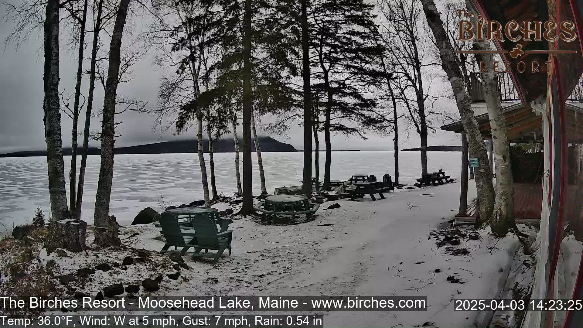 A snow-covered lakeside area with picnic tables and chairs, bare trees, and a partially frozen lake under a cloudy sky.