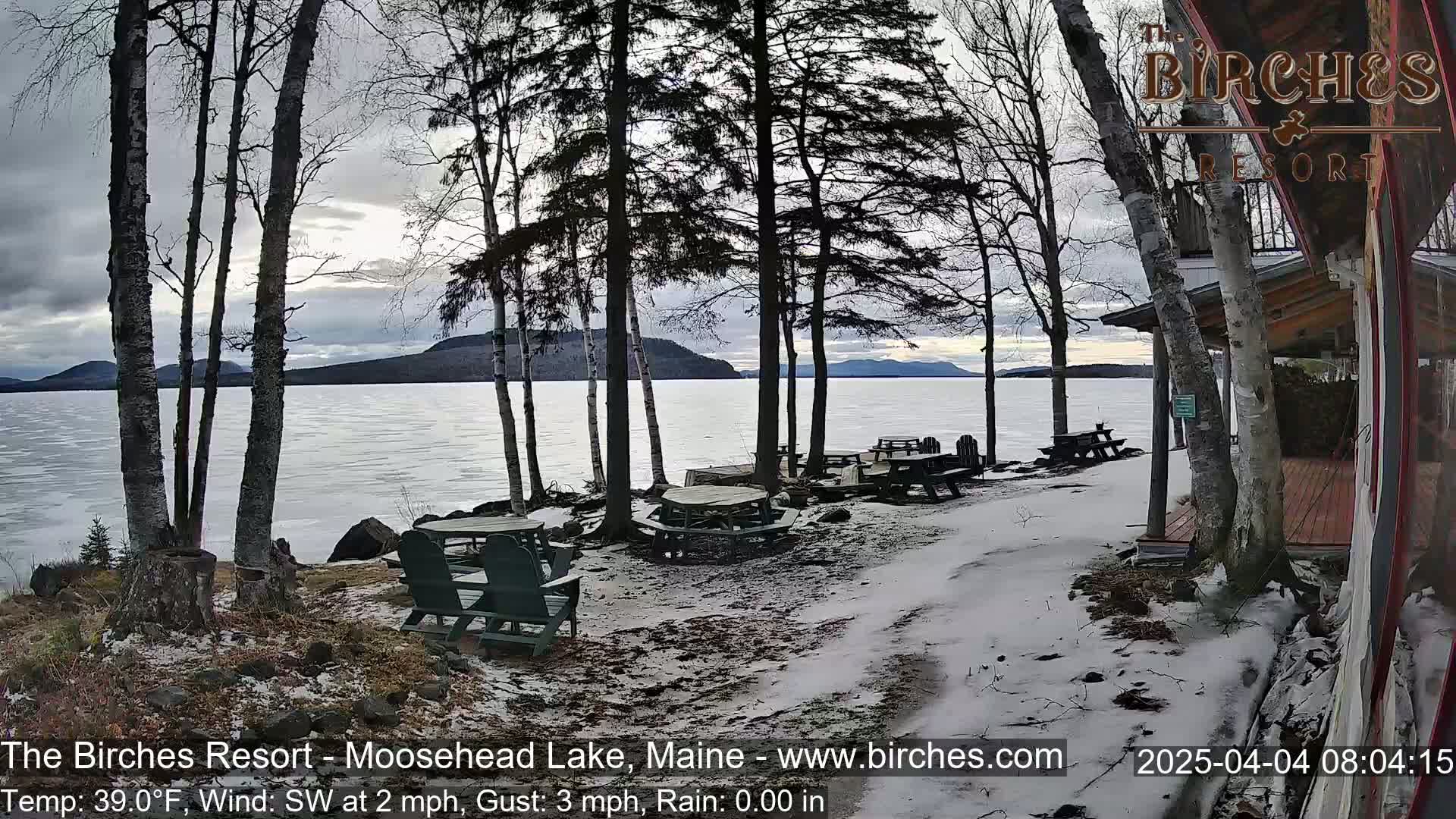 A partly cloudy, cool day shows a snow-dusted lakeside patio with empty picnic tables and chairs overlooking a partially frozen lake and distant mountains.