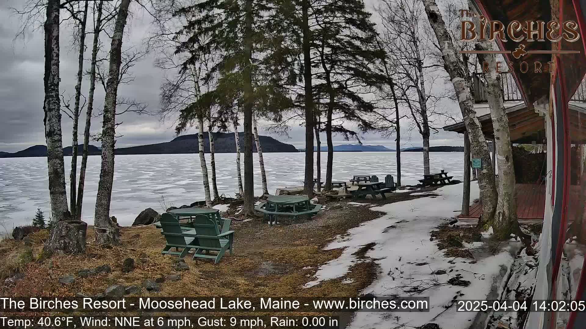 A partially frozen lake is visible beyond several picnic tables and chairs situated on a snow-dusted lakeside area under an overcast sky.