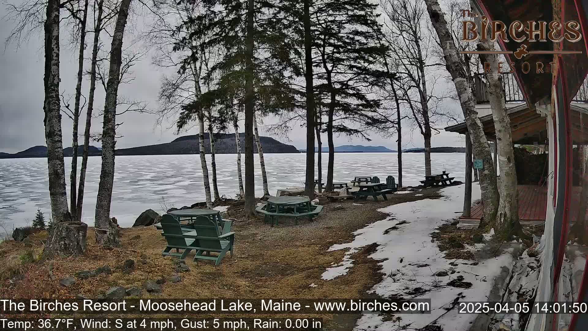 A partially frozen lake is viewed from a lakeside resort with picnic tables and chairs, under a cloudy sky.