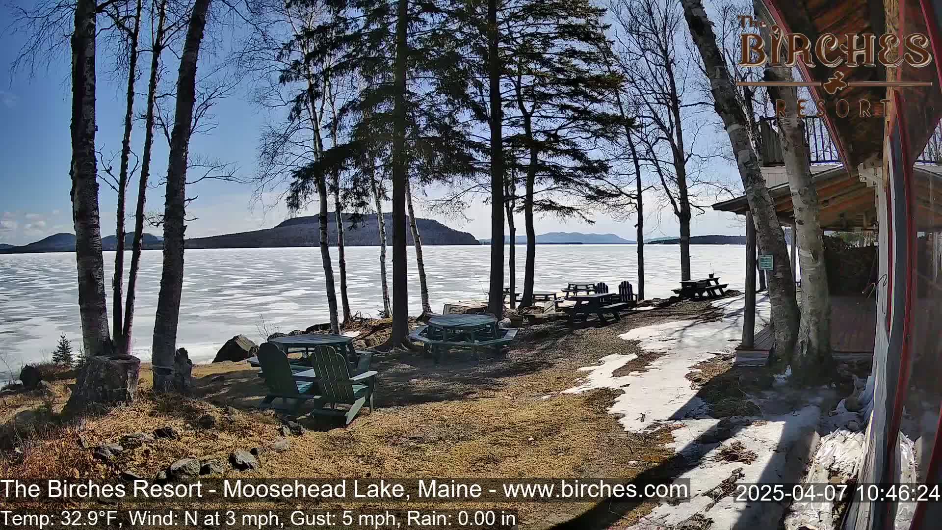 A partly sunny day reveals a partially frozen lake viewed from a lakeside patio with picnic tables and surrounding trees, some patches of snow remain on the ground.