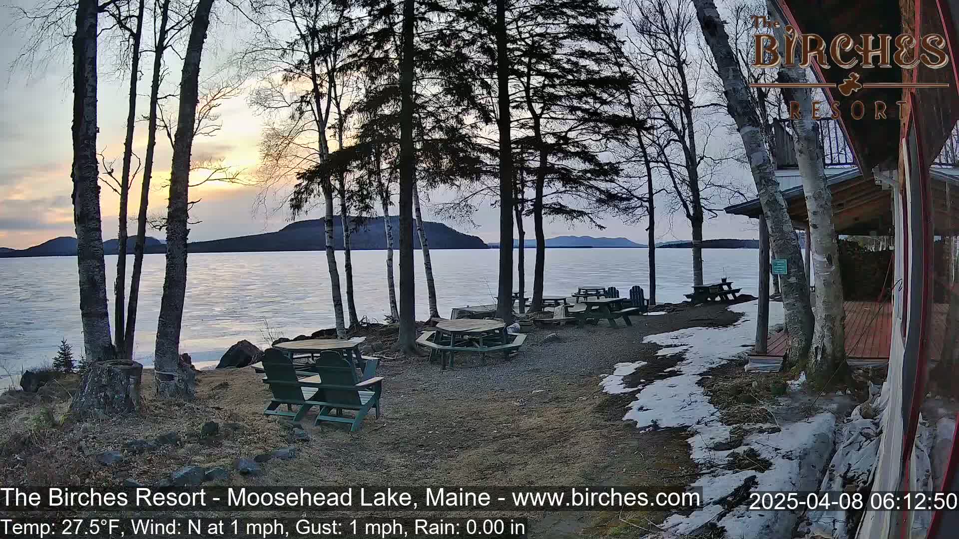 A lakeside patio with picnic tables and chairs, partially covered in snow, overlooks a partially frozen lake at sunset under a partly cloudy sky.
