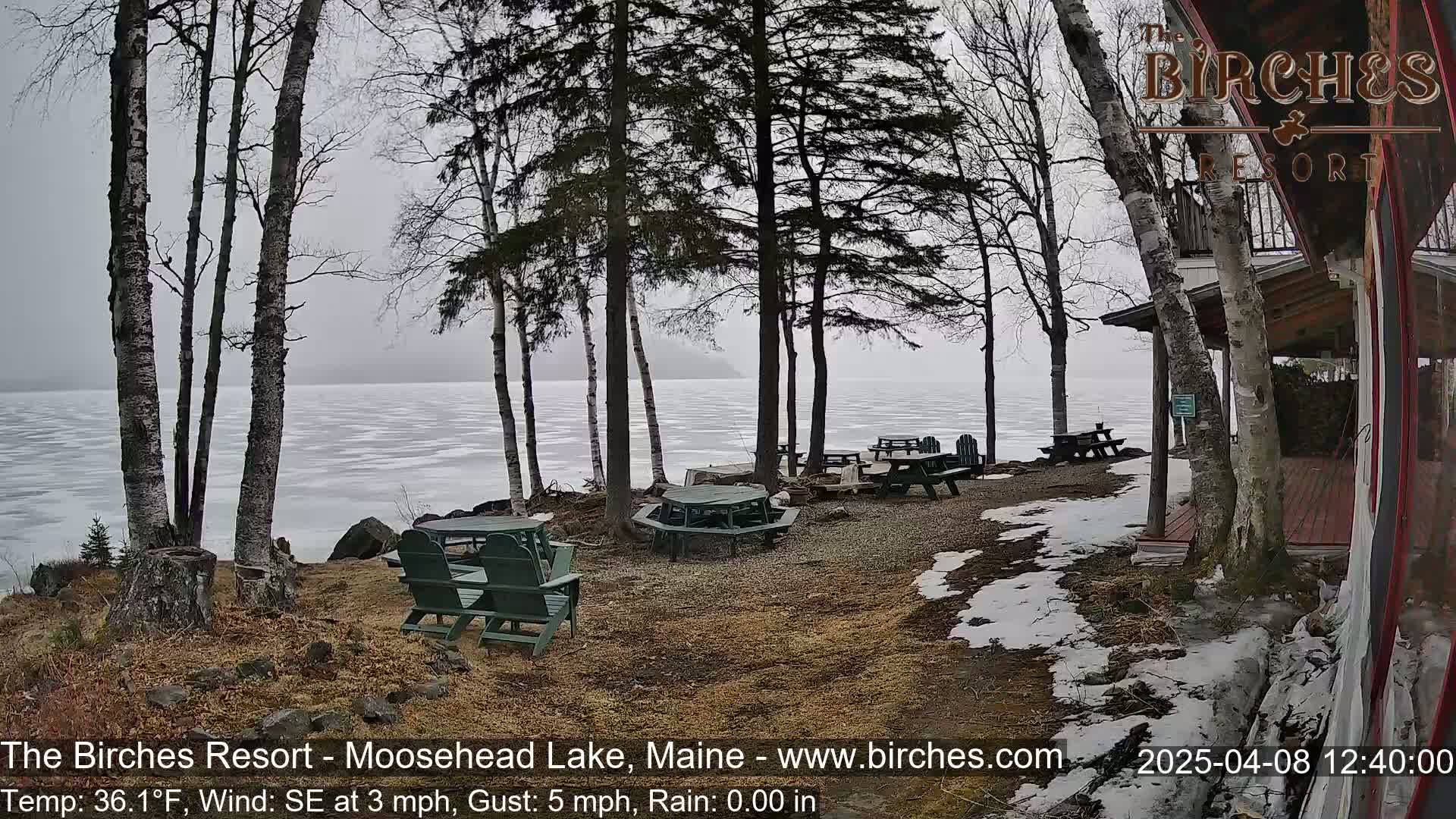 A lakeside resort area with several empty picnic tables and chairs, partially covered in snow, overlooks a partially frozen lake under an overcast sky.