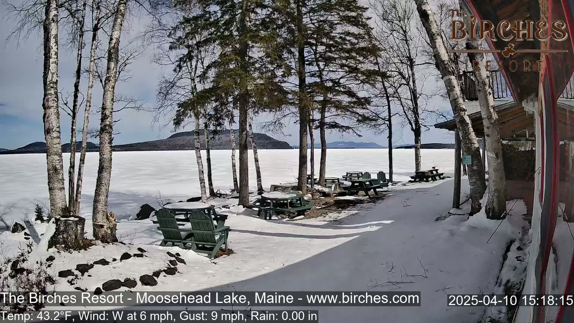 A snow-covered lakeside patio with empty picnic tables and chairs, under a partly sunny sky, overlooks a frozen lake and distant mountains.