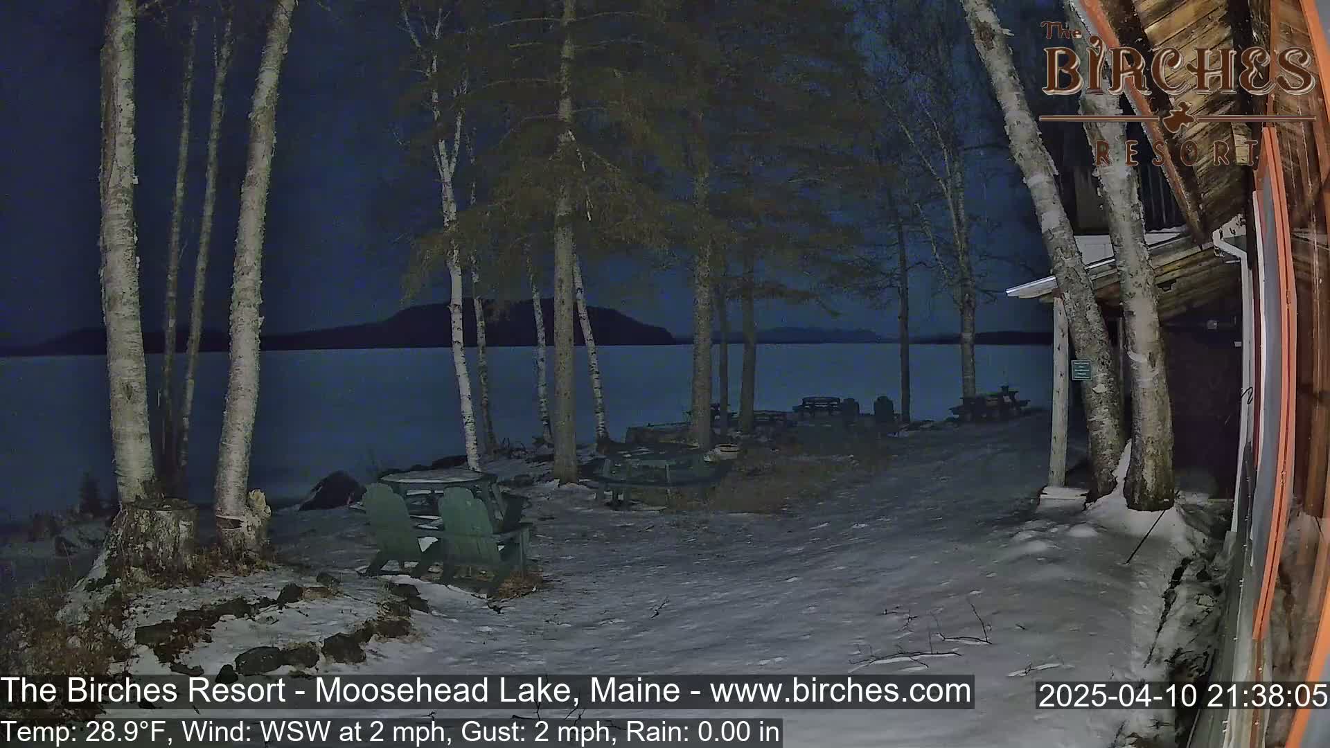 A snow-covered lakeside area at night with several empty picnic tables and chairs under birch trees, next to a building, overlooking a calm lake under a dark sky.