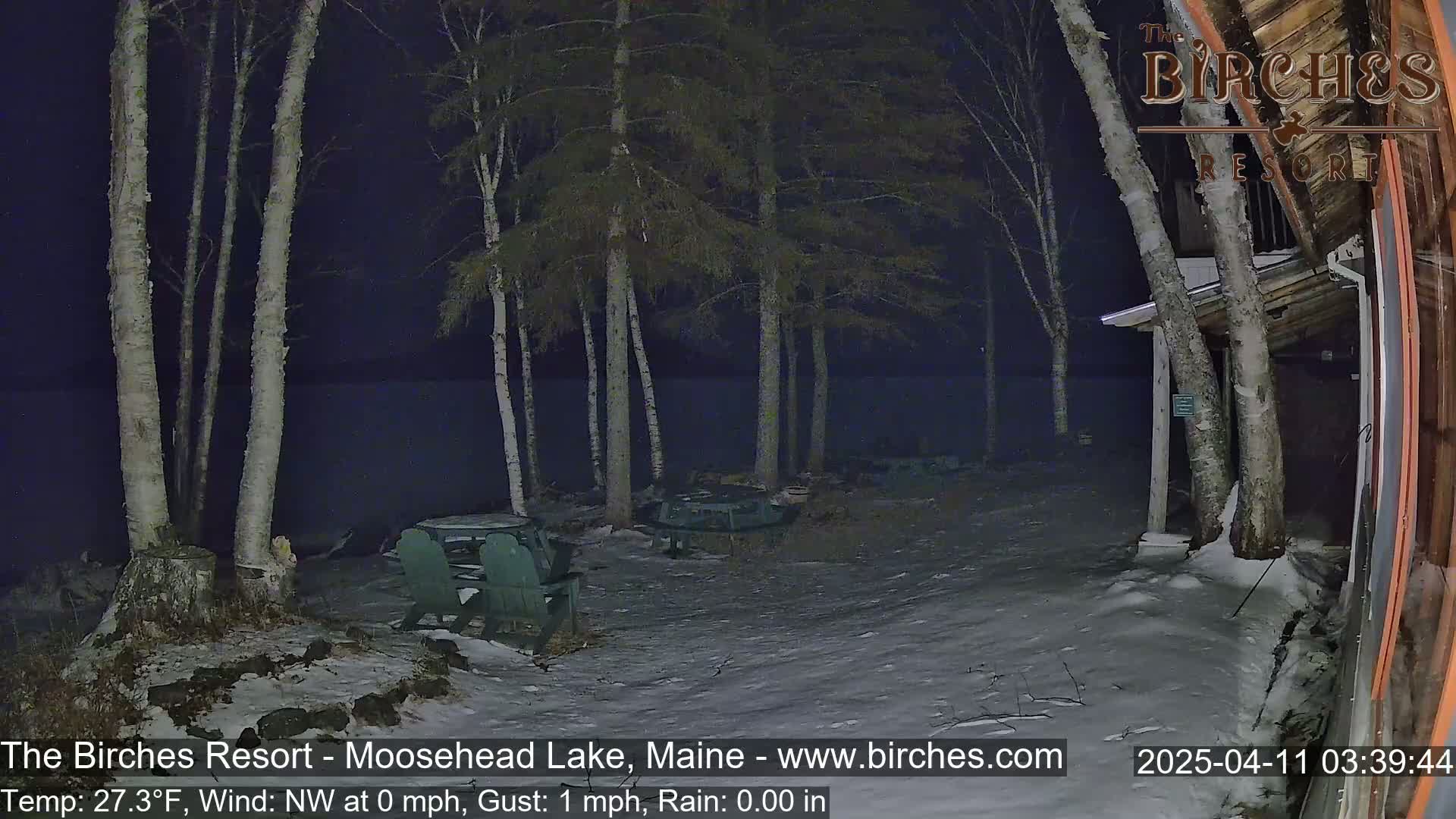A snow-covered lakeside patio at night, illuminated by artificial light, features several empty Adirondack chairs and tables near a building, with birch trees in the background under a dark sky.