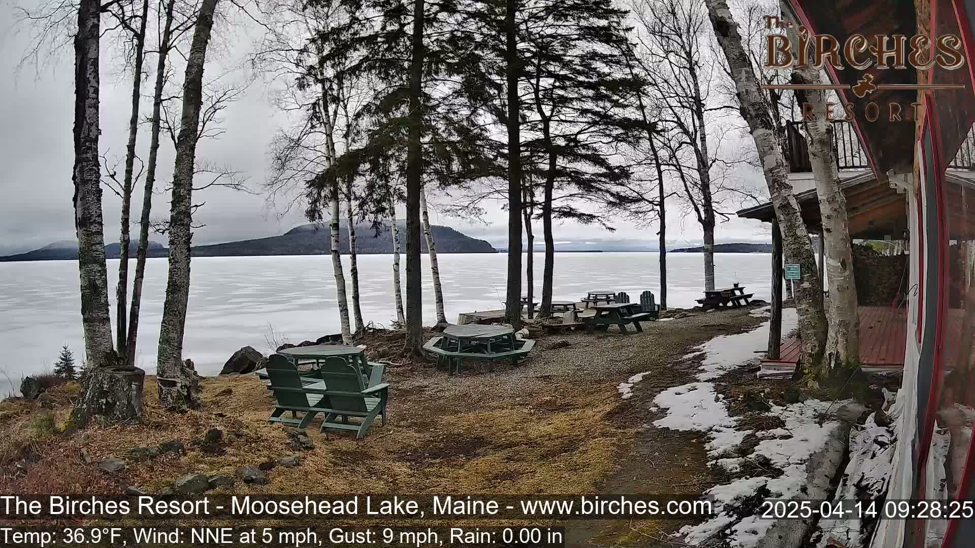 A partially frozen lake is visible beyond a lakeside area with several picnic tables and chairs, under a gray, overcast sky.