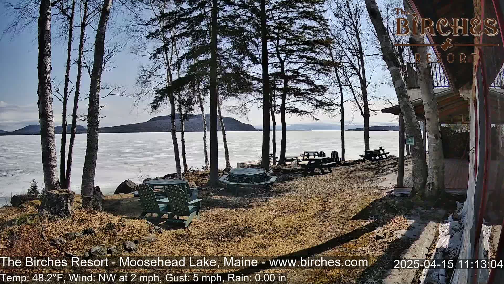 A partly frozen lake is visible beyond a lakeside patio area with picnic tables and chairs under a mostly sunny sky.