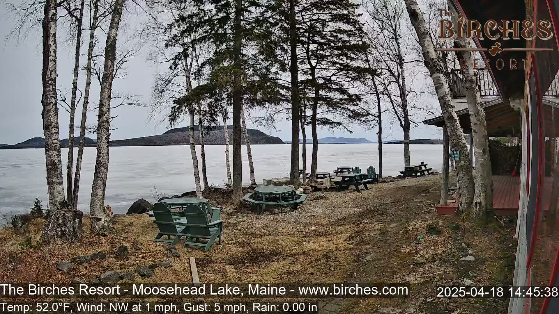 A lakeside patio with several empty picnic tables and chairs, overlooking a partly frozen lake under an overcast sky.
