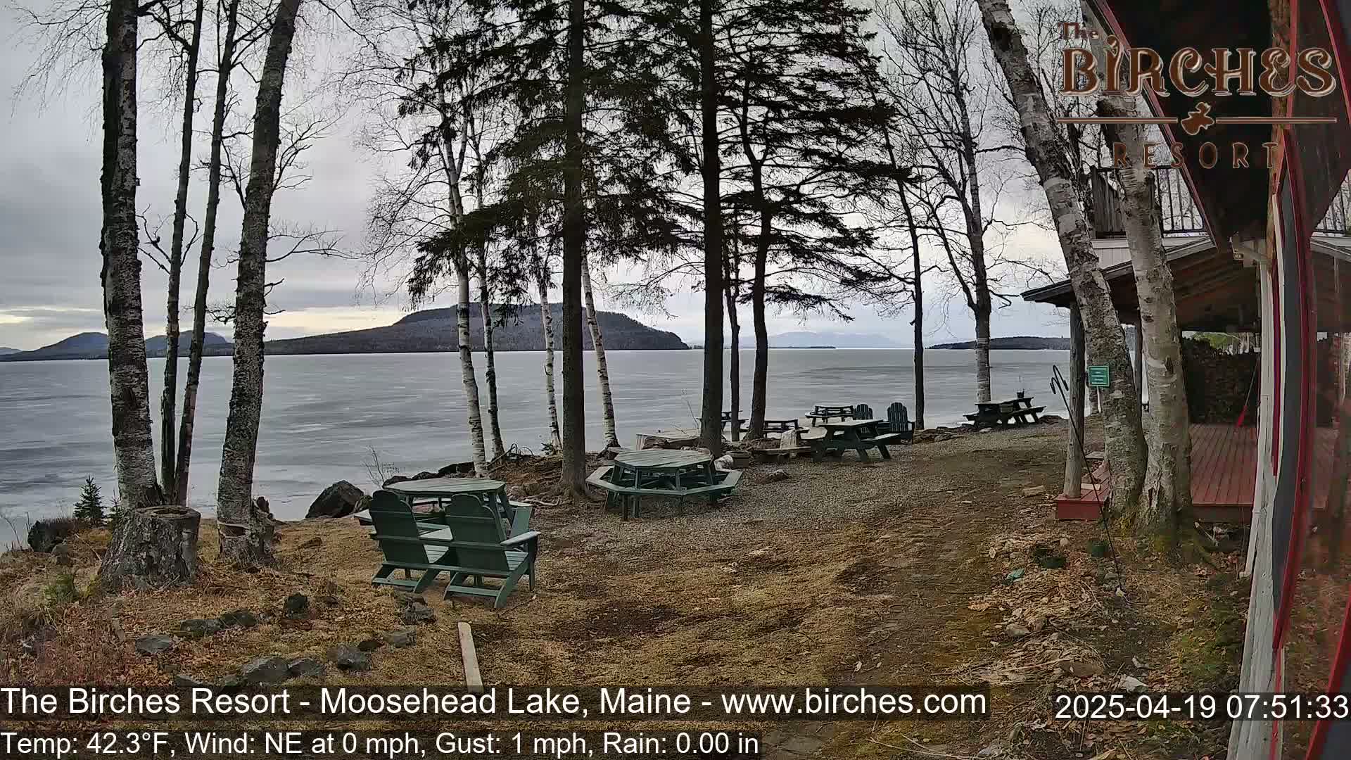 A partially frozen lake is visible beyond a lakeside area with picnic tables and chairs under a cloudy sky.
