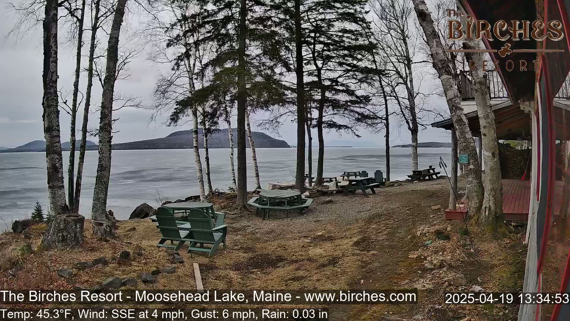 A partly cloudy day reveals a lake partially covered in ice, with picnic tables and chairs situated on a lakeside patio near bare trees and a building.