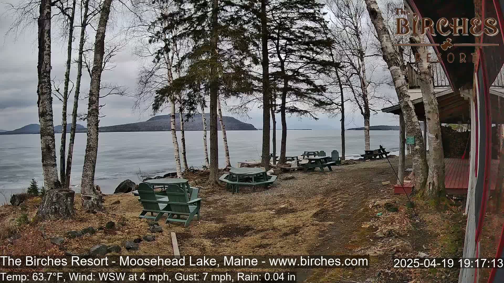 A lakeside patio with picnic tables and chairs is partially visible under a gray sky with light rain, overlooking a partly frozen lake and distant mountains.