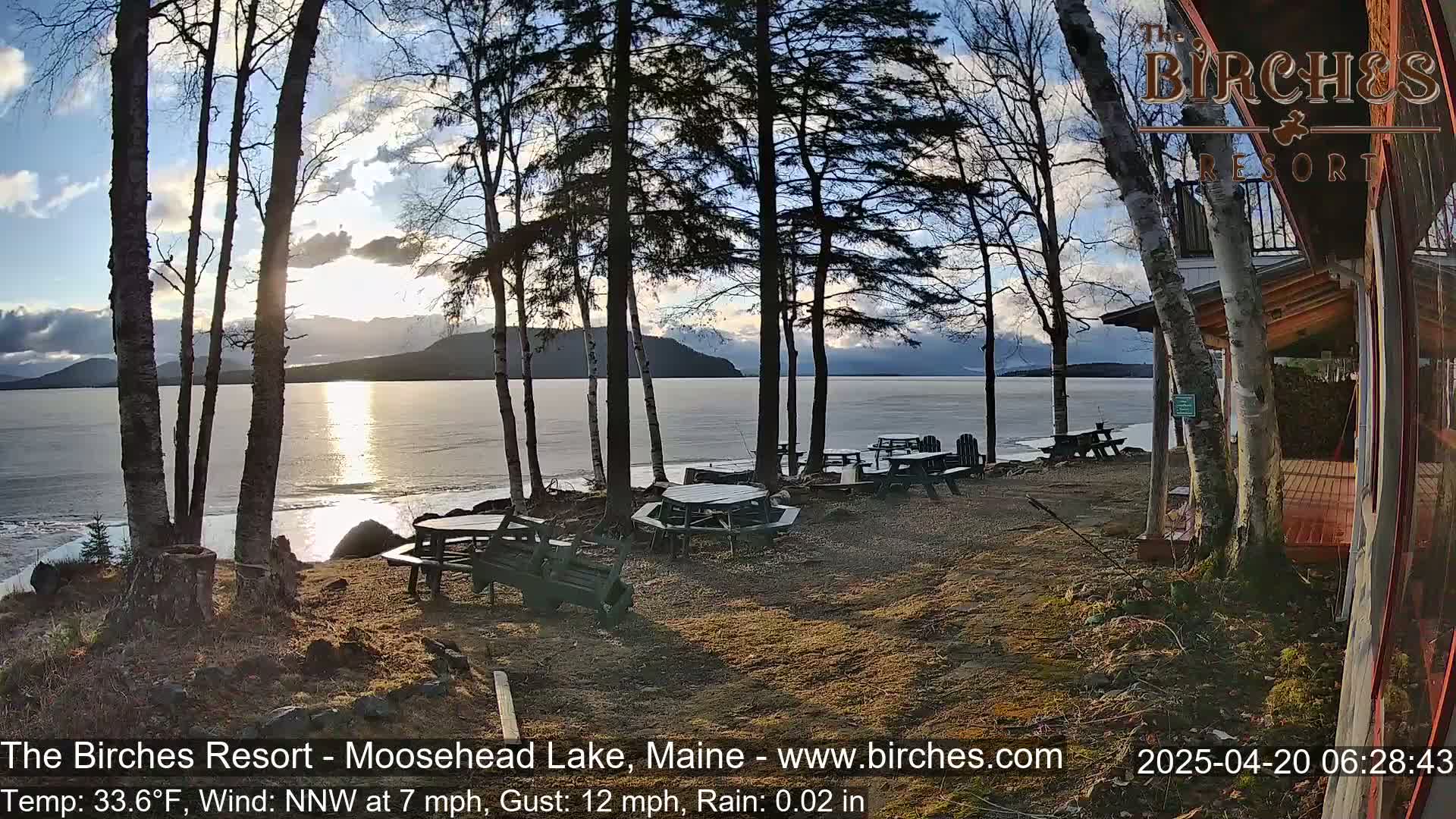 Several picnic tables sit near a lakeside building under a partly sunny sky with a light wind and rain.