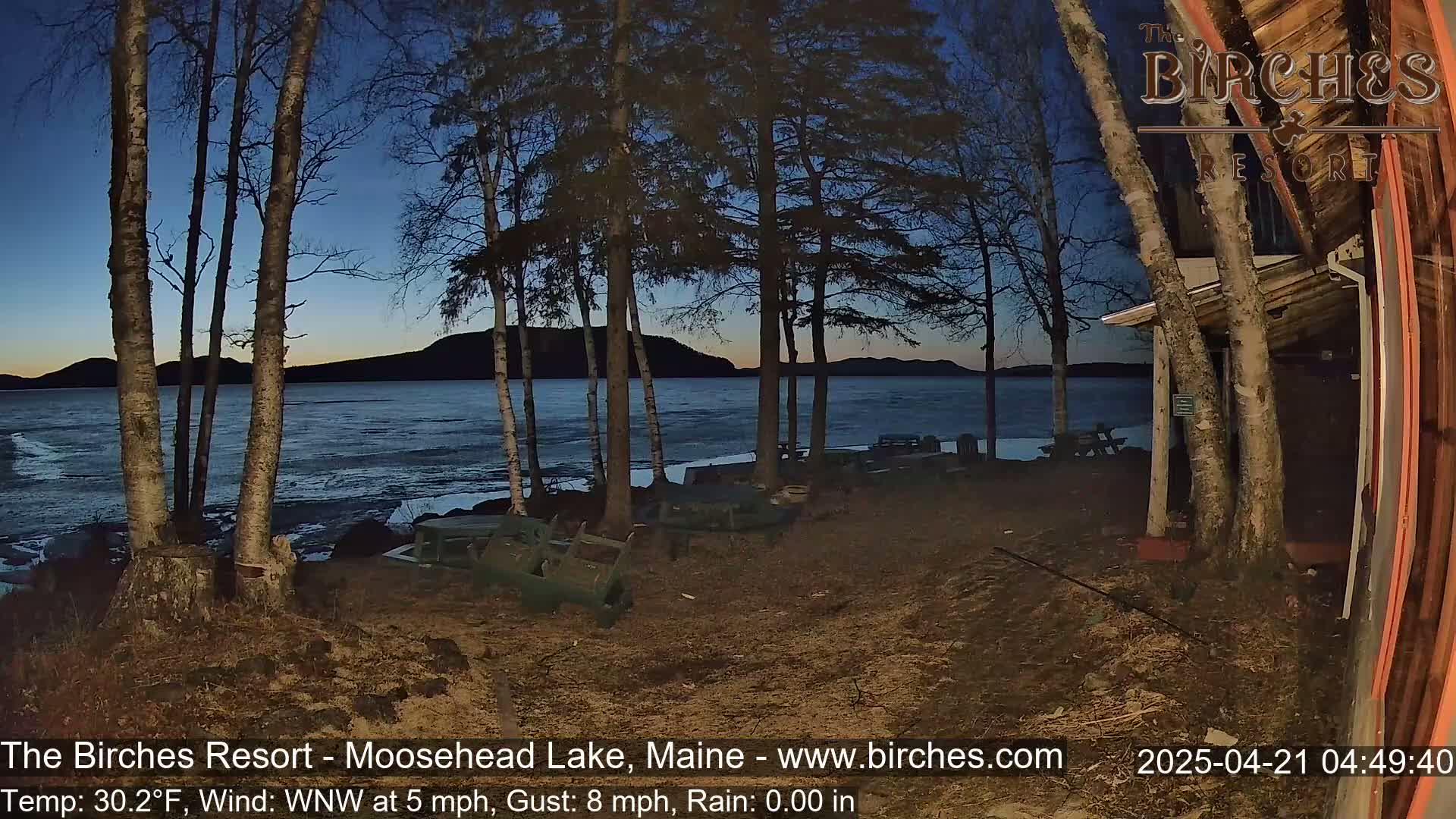 A twilight view of a lake with partially frozen shoreline, viewed from a lakeside property with bare trees, some picnic tables, and scattered debris on the ground, under a clear, dark blue sky.