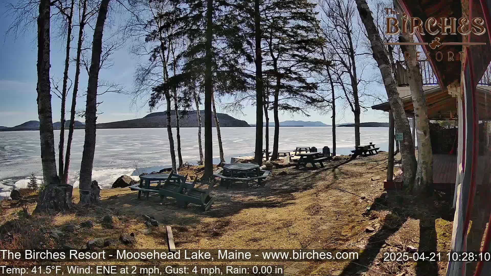 A lakeside picnic area with several empty tables and chairs is partially shaded by bare trees on a sunny day with a partly frozen lake in the background.