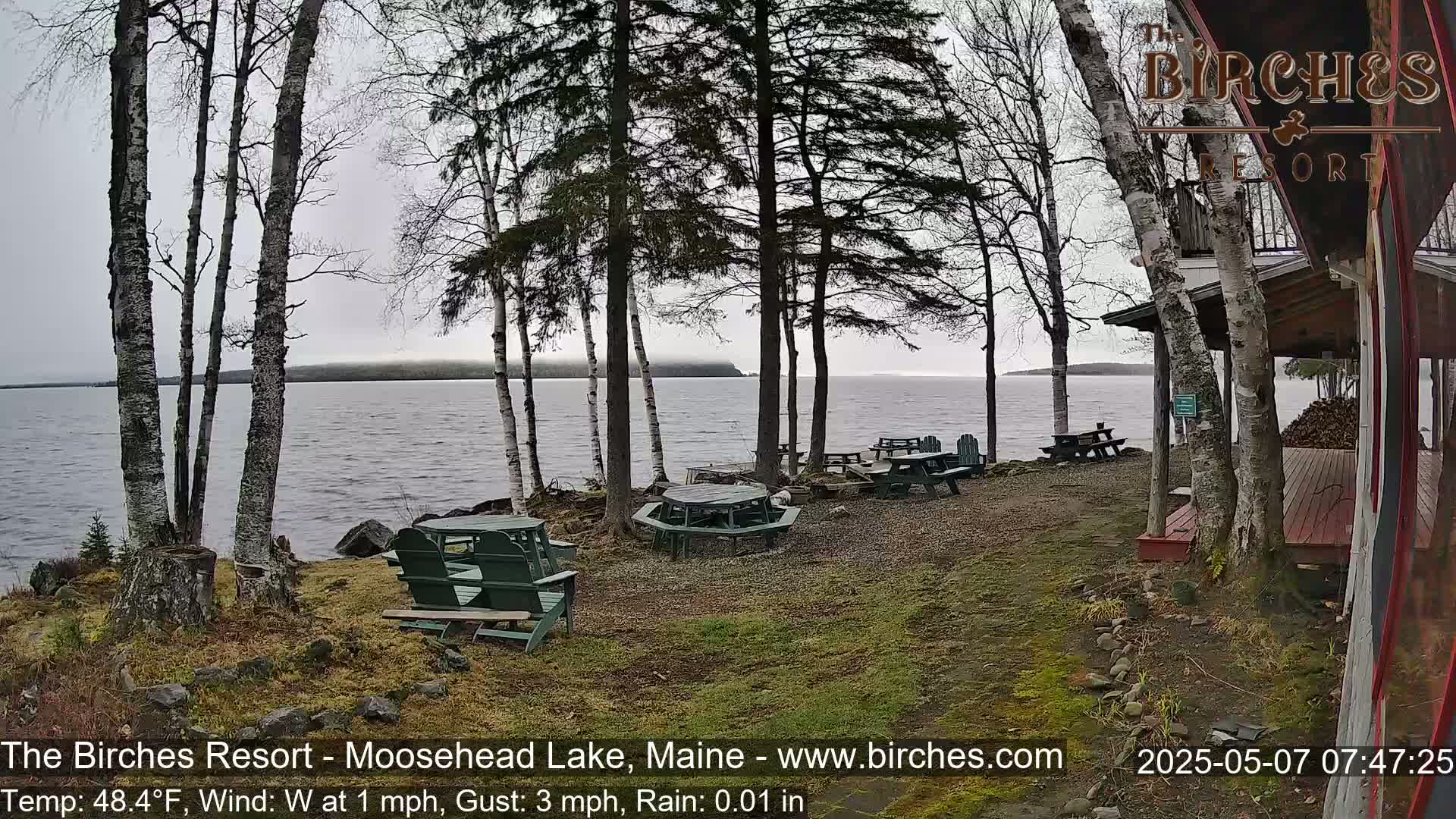 A lakeside patio with picnic tables and chairs under a gray, rainy sky.