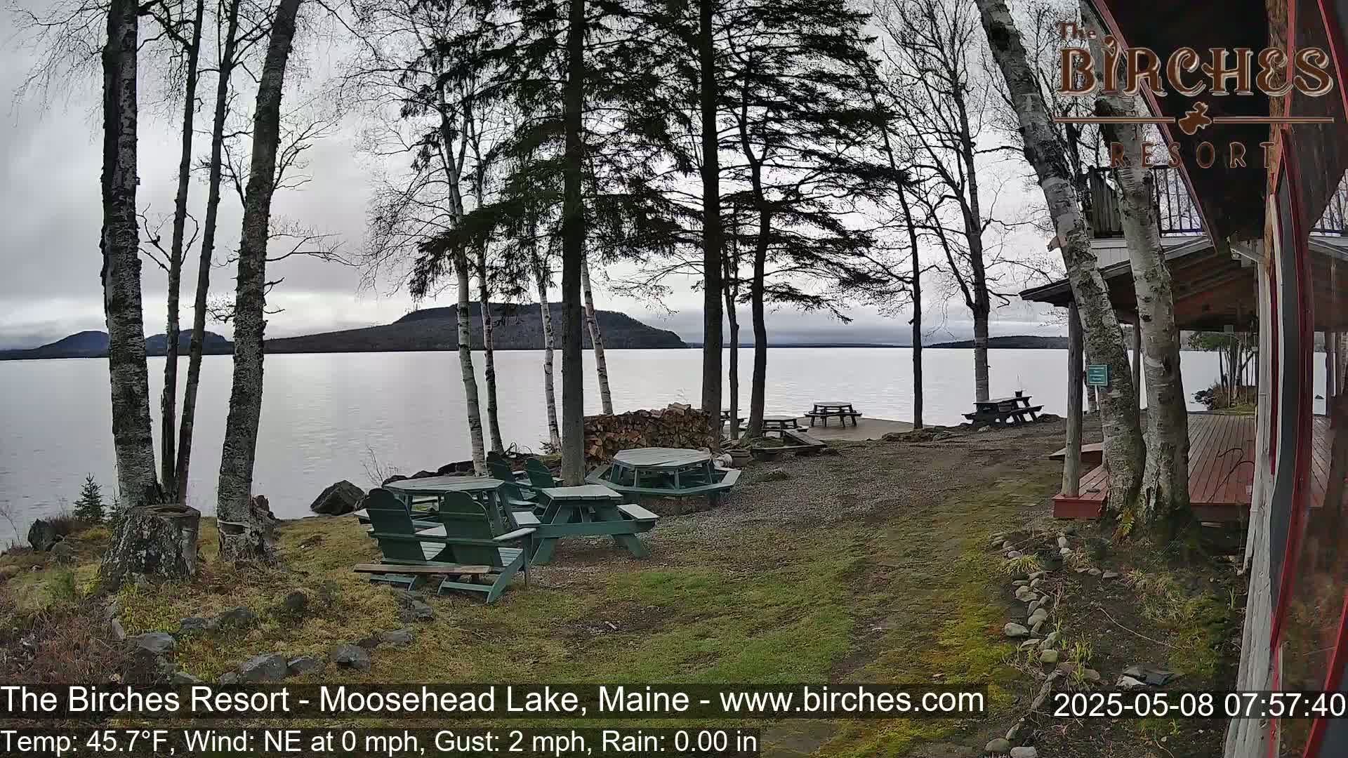 Overlooking a calm lake, several picnic tables sit on a grassy area next to a lakeside building under a cloudy sky.