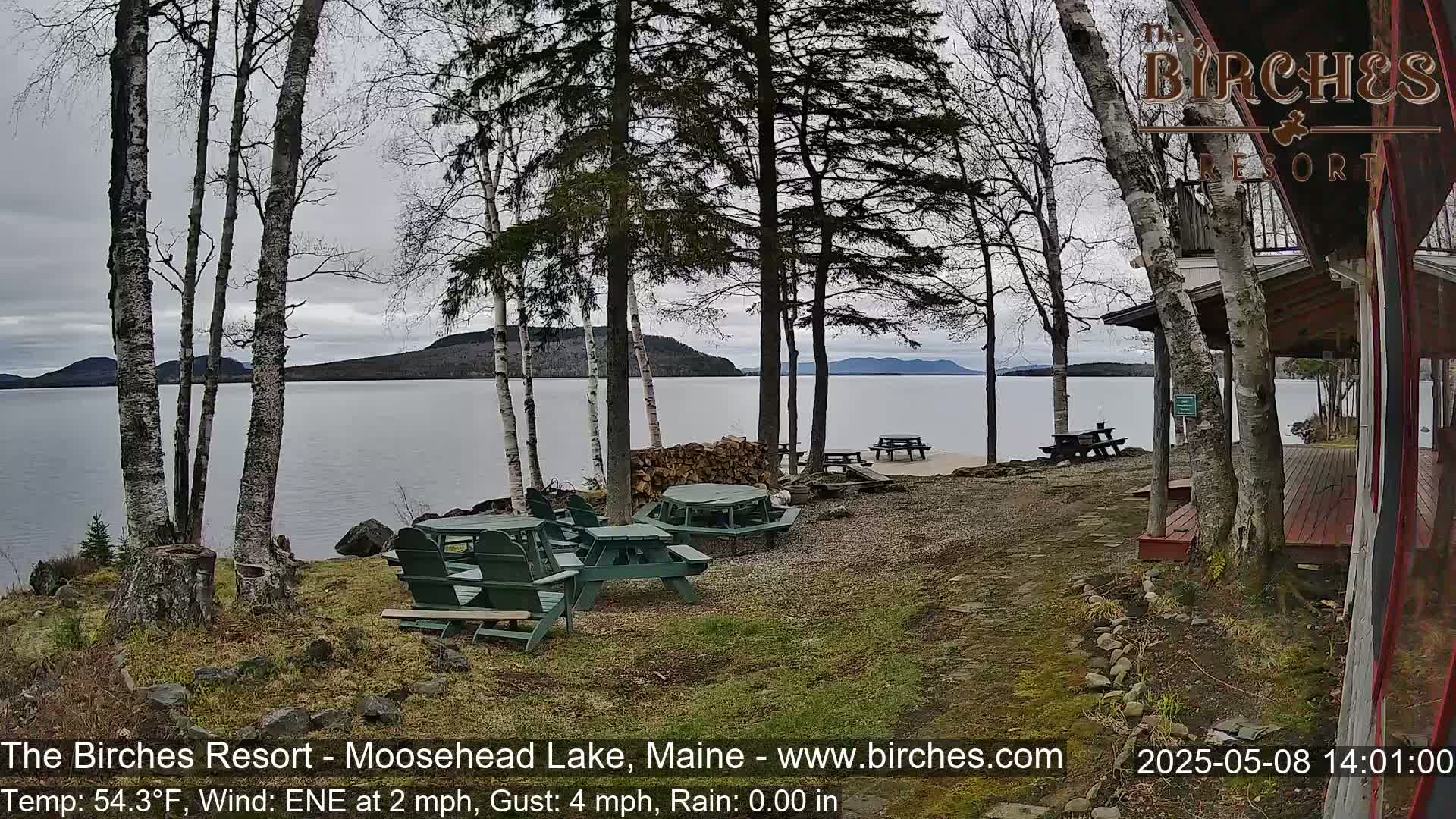 A lakeside view shows several green picnic tables and chairs near a cabin under a cloudy sky.